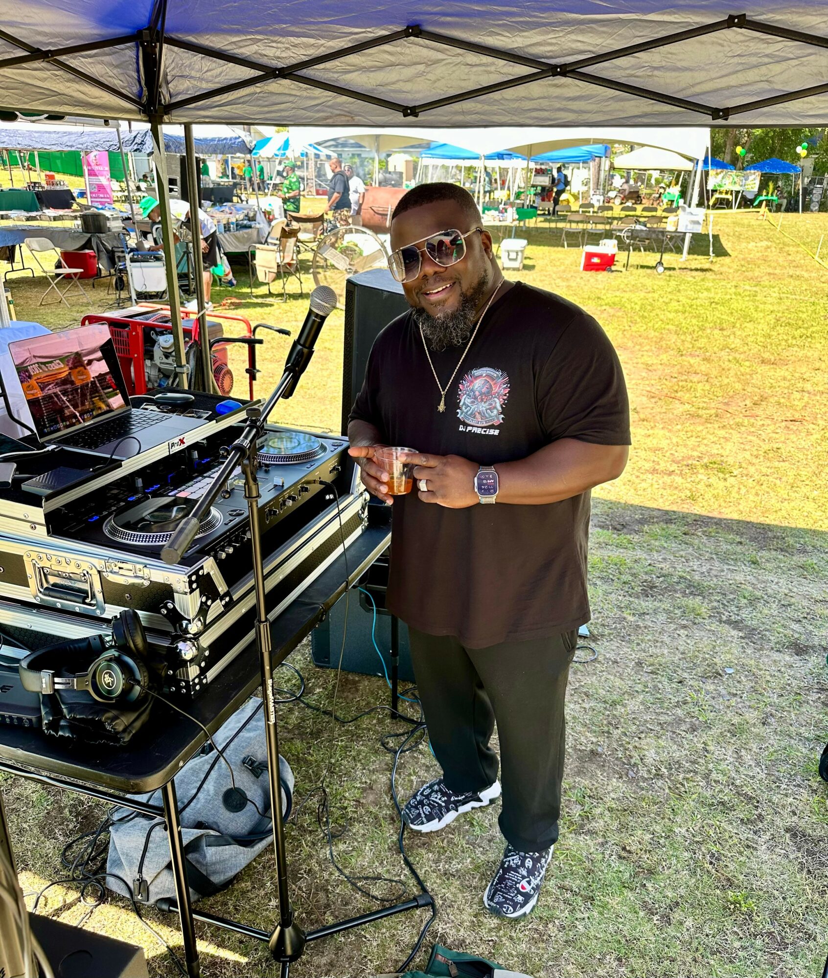 Man wearing sunglasses and a black t-shirt stands under a canopy with DJ equipment, holding a drink, outdoors on grass.