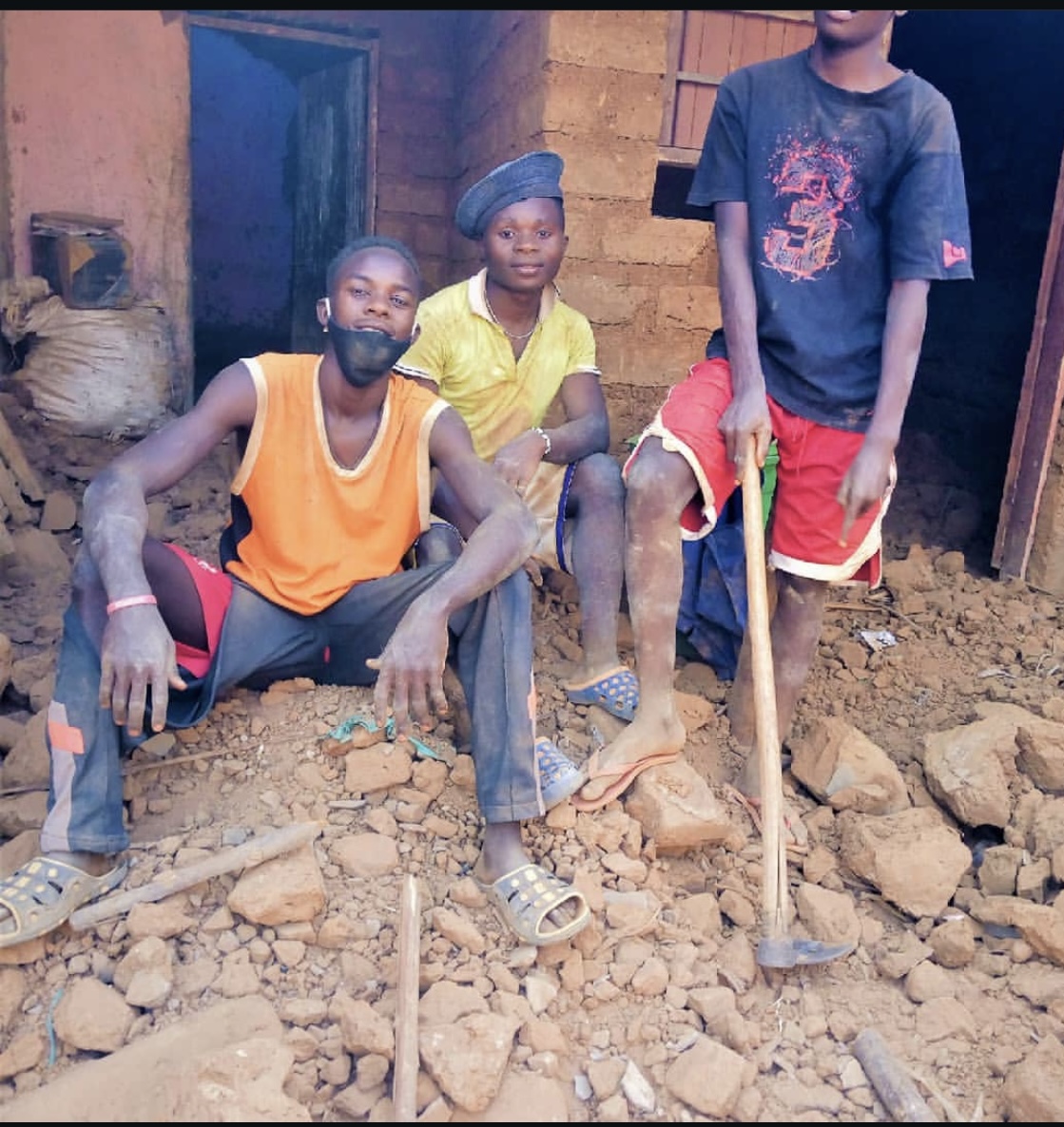 Three children standing and sitting on rocky ground outside a building, one holding a stick, with a door and wall in background.