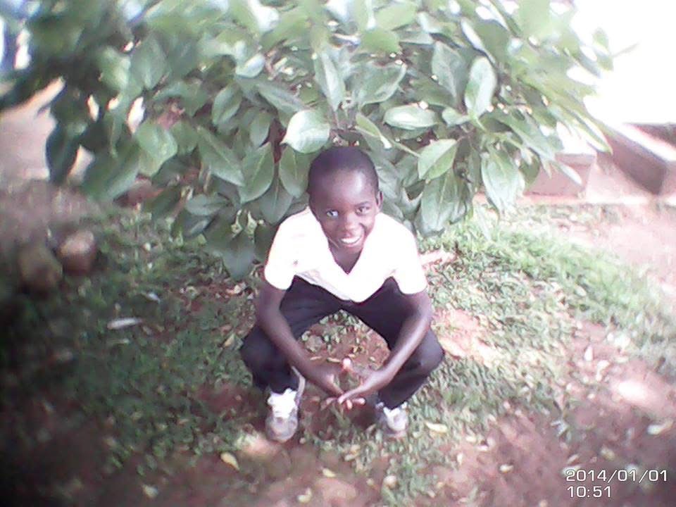 Child crouching under a large leafy tree, smiling, outdoors on grass and dirt, wearing a white shirt and dark shorts.