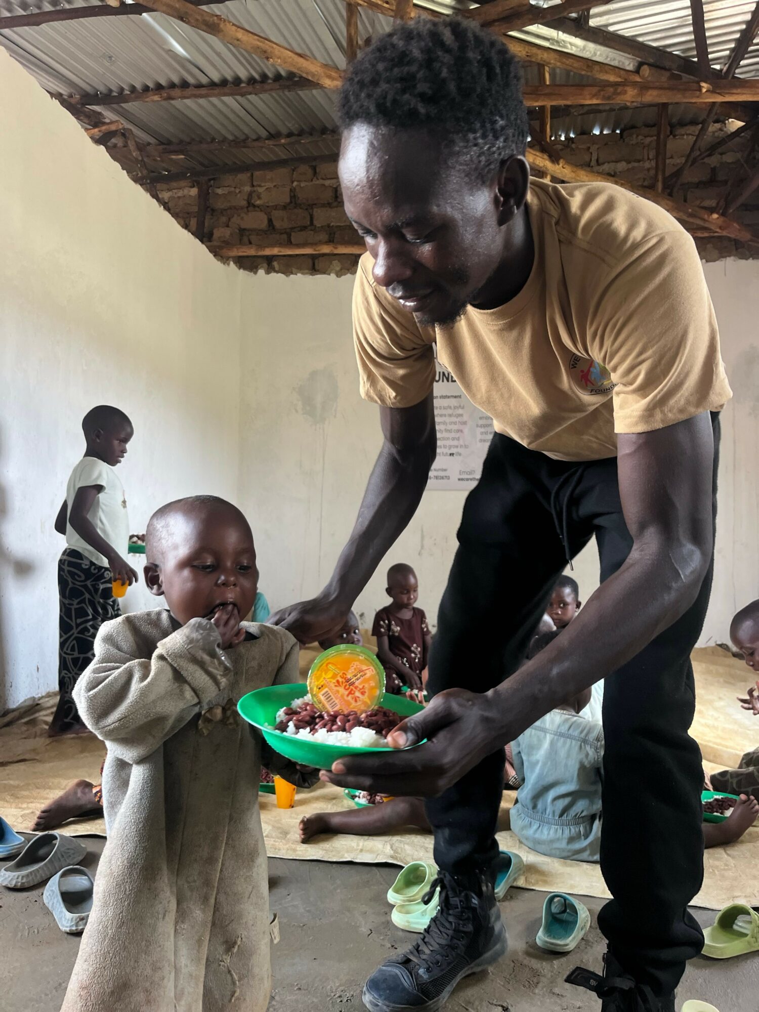 Man handing a colorful object to a young child in a rustic setting with other children nearby.