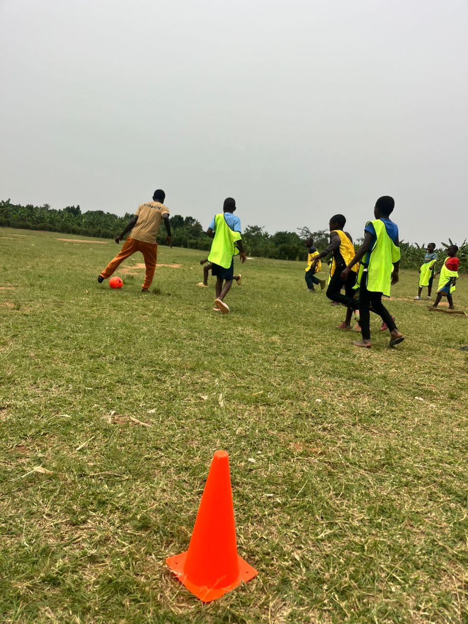 Children playing soccer on a grassy field, some wearing yellow vests, with an orange cone in the foreground, overcast sky.