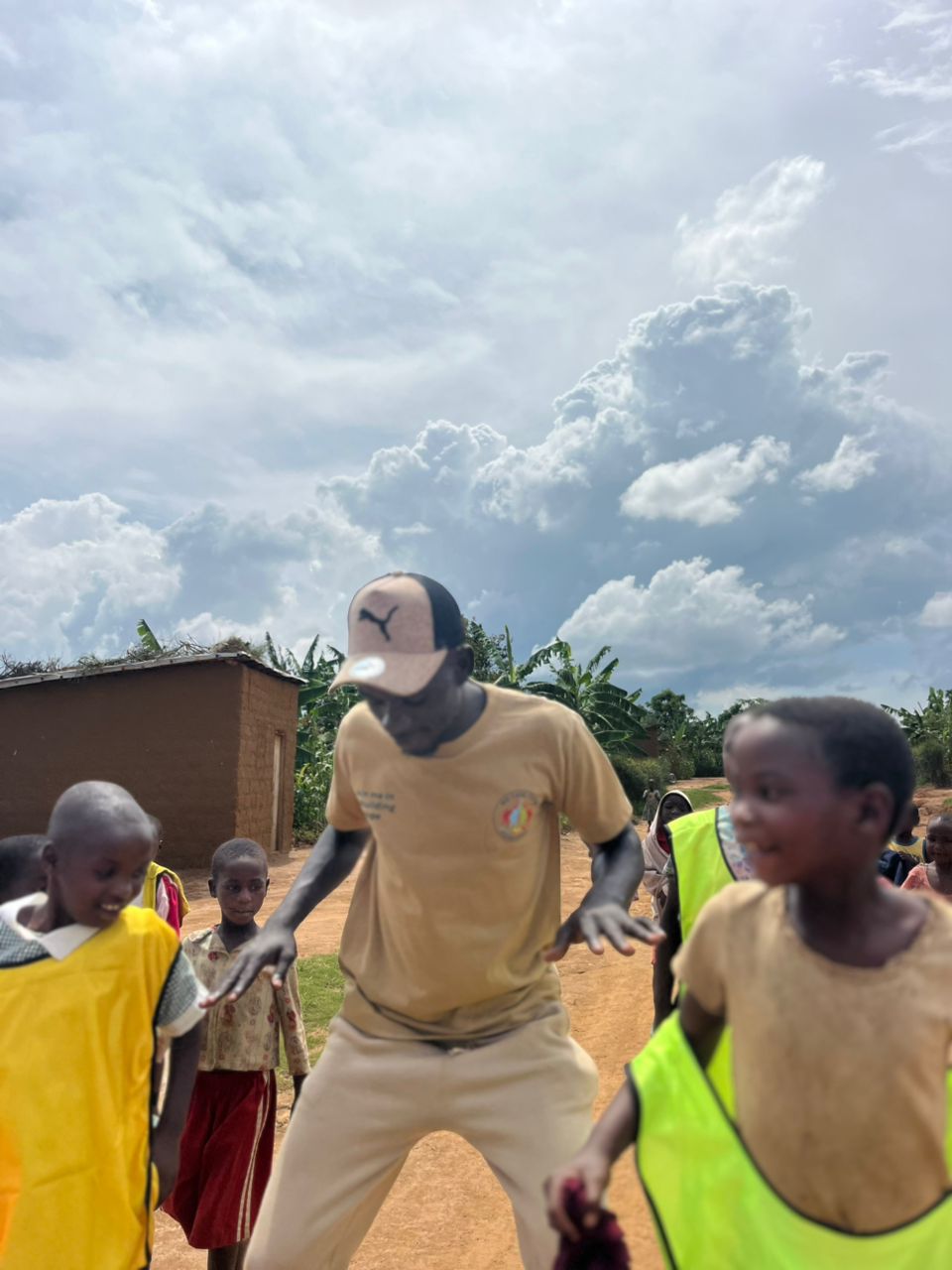 Man in beige clothing and cap interacts with children outdoors under cloudy sky, some children wearing yellow vests.