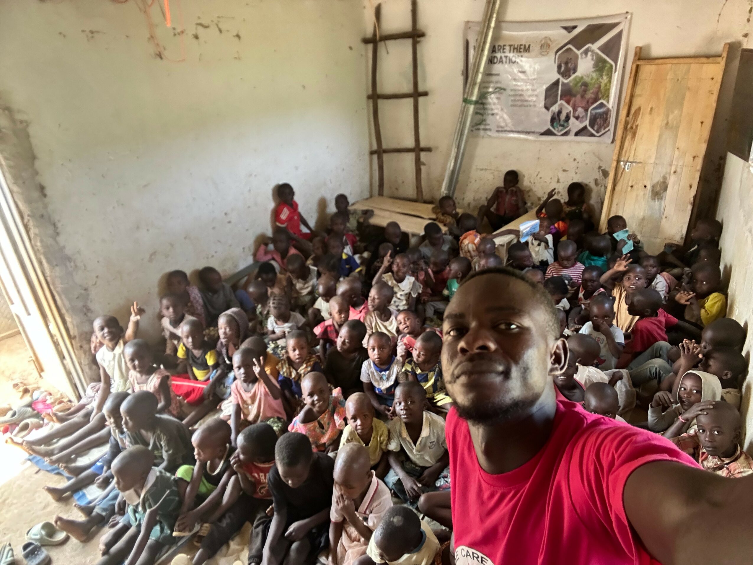 A man taking a selfie with a large group of children sitting on the floor inside a room with unfinished walls and wooden structures.