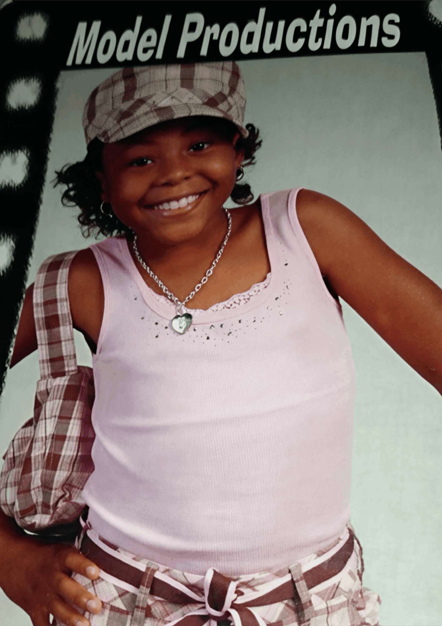Smiling girl wearing a hat, pink top, and plaid accessories, standing against a white background.