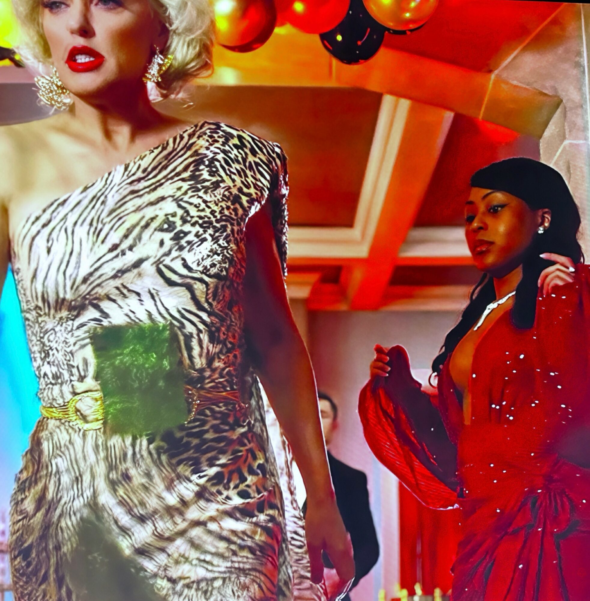 Two women in glamorous outfits at an indoor event, with colorful balloons overhead and a decorated ceiling.