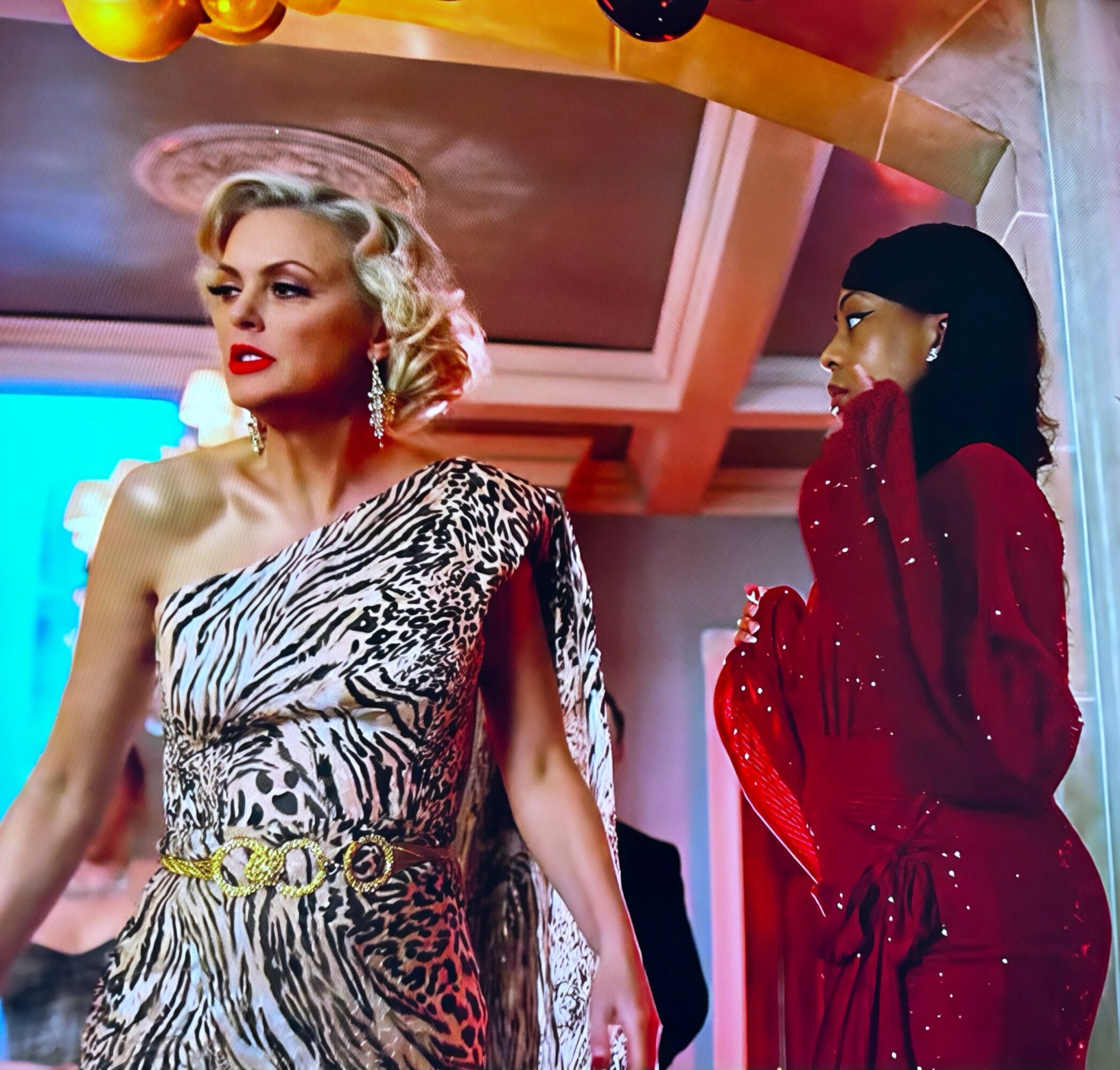 Two women in glamorous dresses at a social event, one in a leopard print gown, the other in a red dress, indoors.