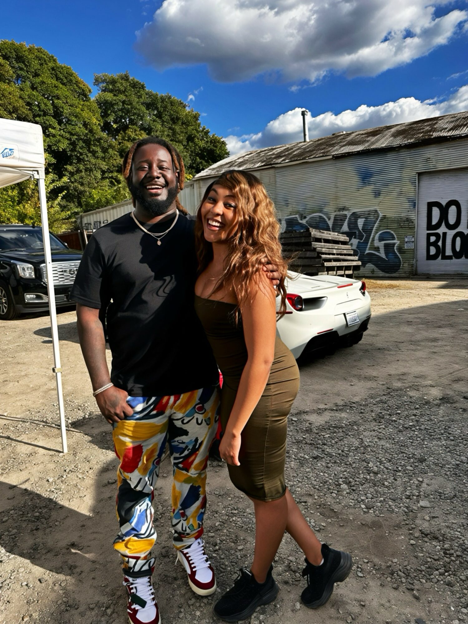 Two people smiling outdoors, standing close together, with a building and parked cars in the background under a partly cloudy sky.