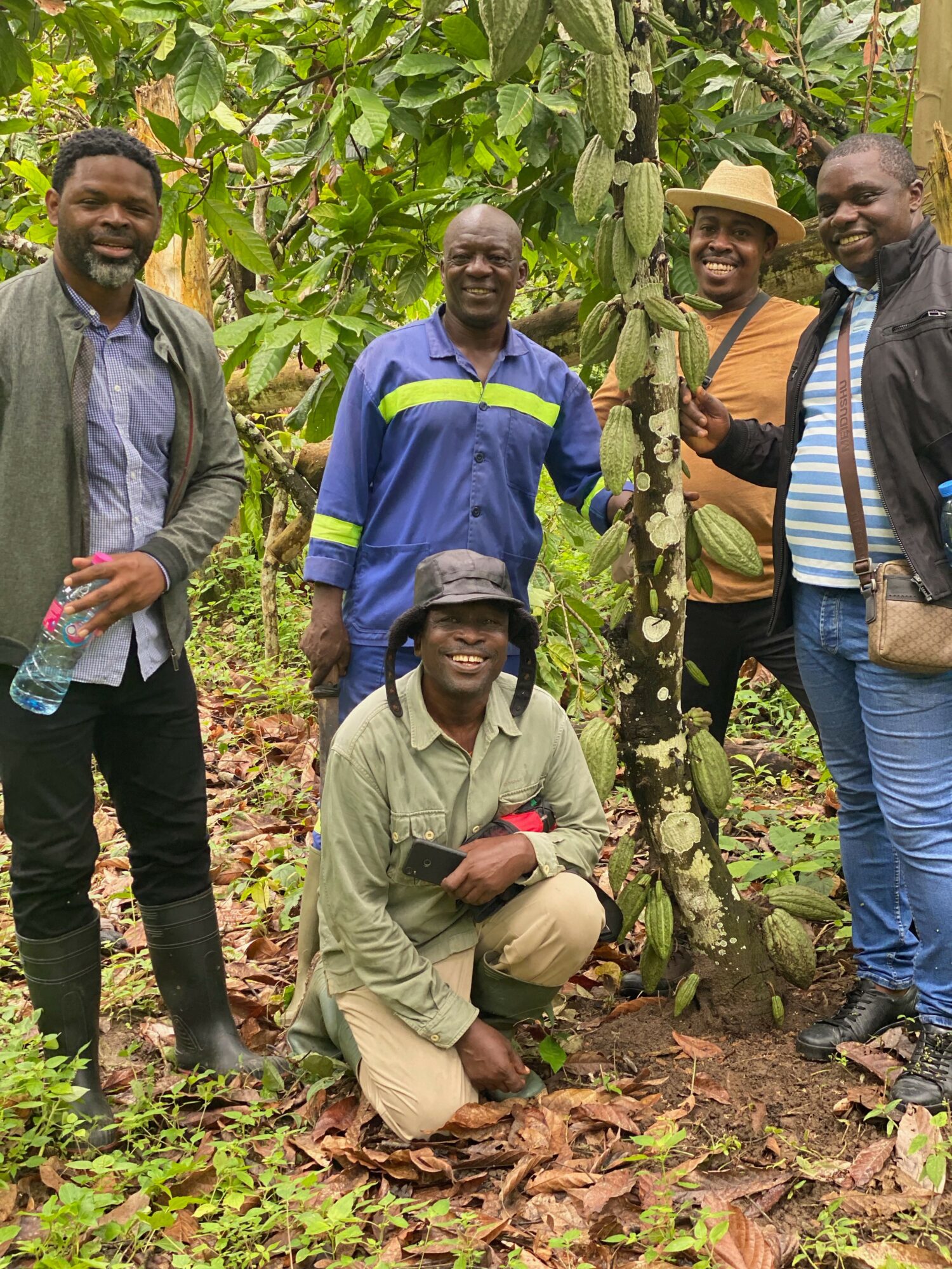 Five people outdoors among green plants, some standing and one kneeling, smiling and holding a water bottle, in a natural setting.