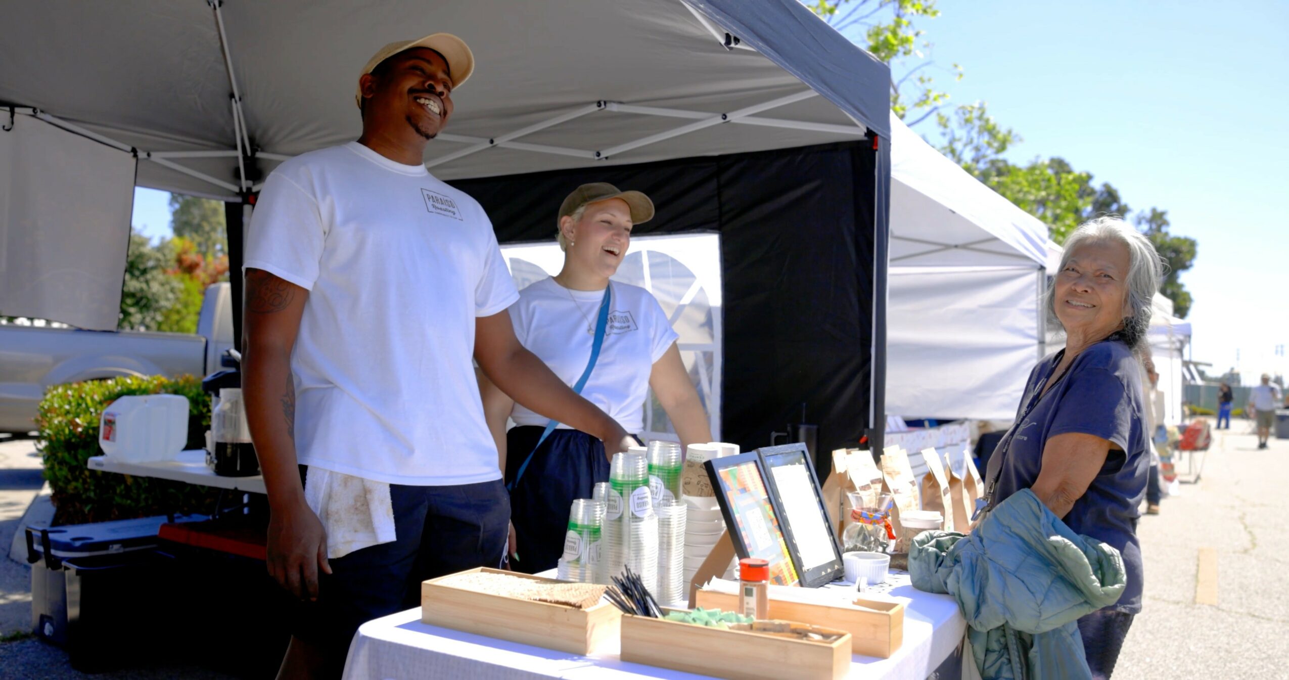 Three people at an outdoor market stall with tents, one person smiling, two others looking at items on display.