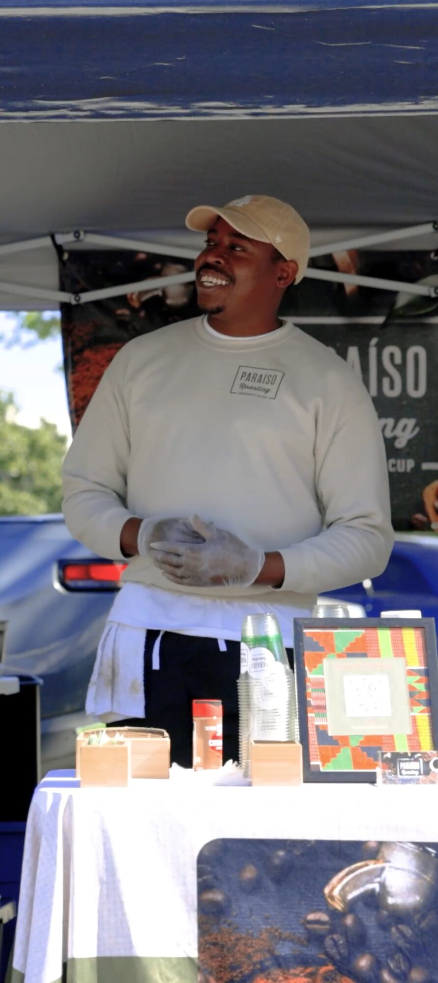 Person wearing a beige cap and white shirt standing behind a table with products, smiling outdoors.