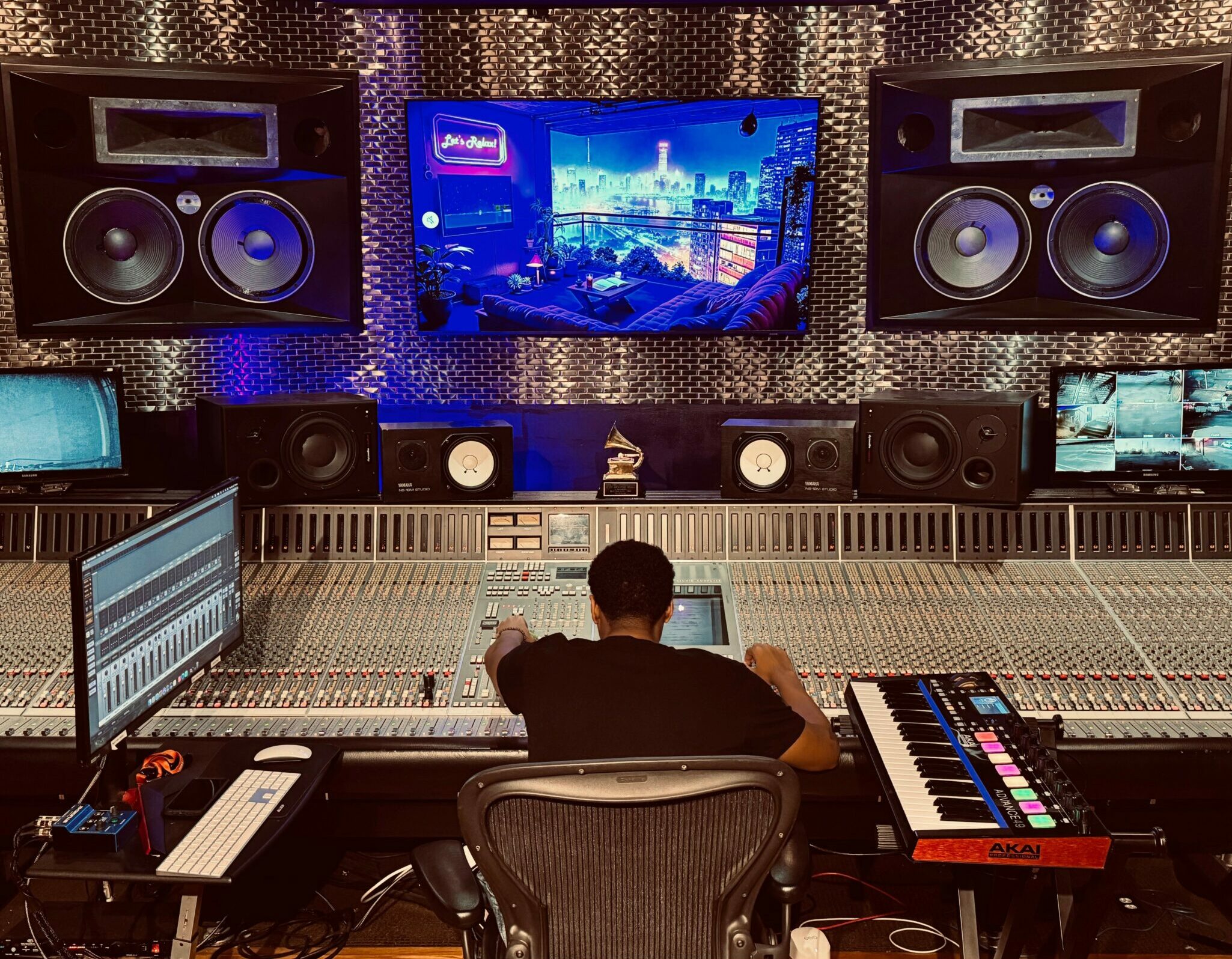 Person working at a music studio mixing console with speakers and a large screen displaying a cityscape.
