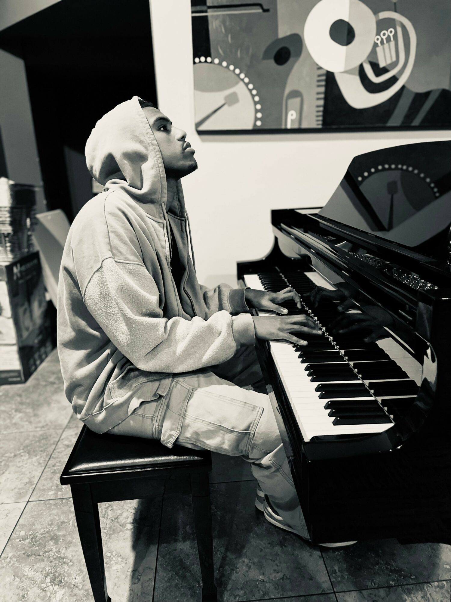 Person wearing hoodie playing piano in a room with abstract wall art.