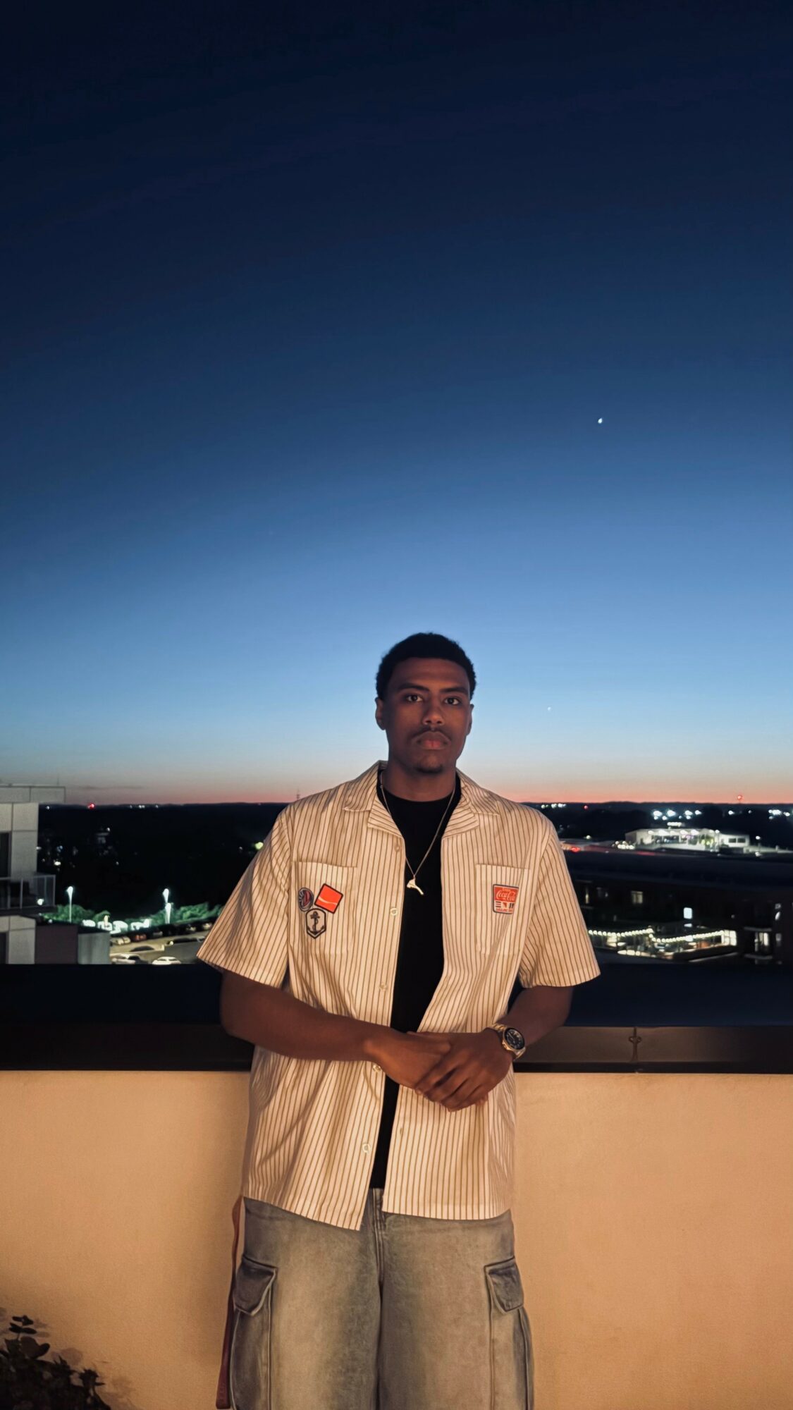 Young man standing outdoors on a balcony during twilight, cityscape in the background, wearing a beige shirt and jeans.