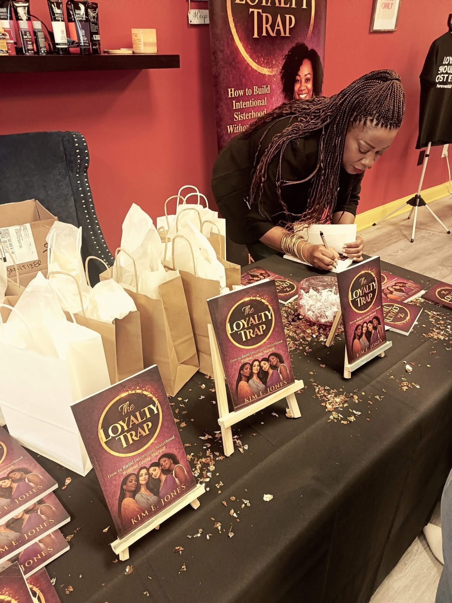 Woman signing books at a table with promotional materials and small signs, in a room with pink walls.