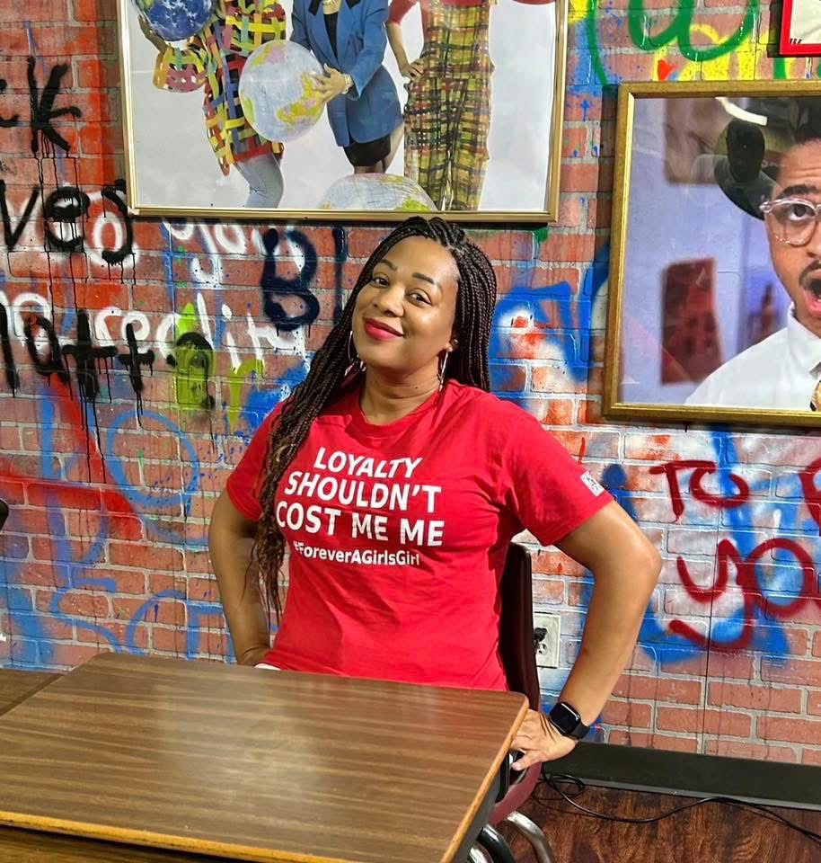 Woman with braided hair wearing a red T-shirt, sitting at a table with graffiti-covered wall behind her.