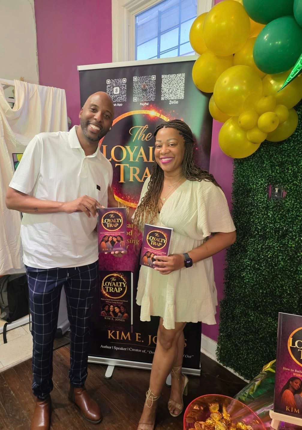 Two people smiling, holding books, standing in front of a promotional banner and balloons, indoors. One person wears a white shirt, the other a light-colored dress.