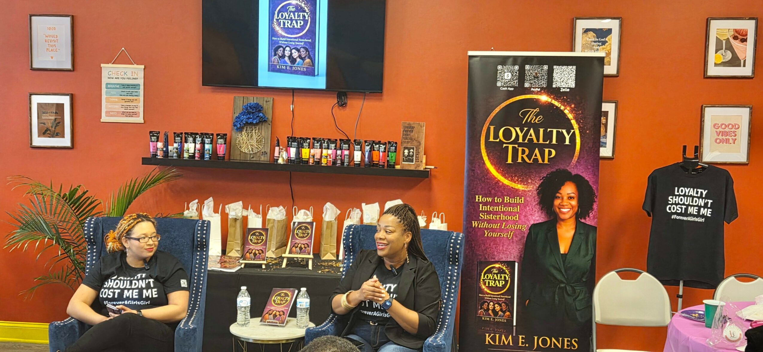 Two women sit in a colorful room with a TV, books, and promotional banners, engaging in conversation.