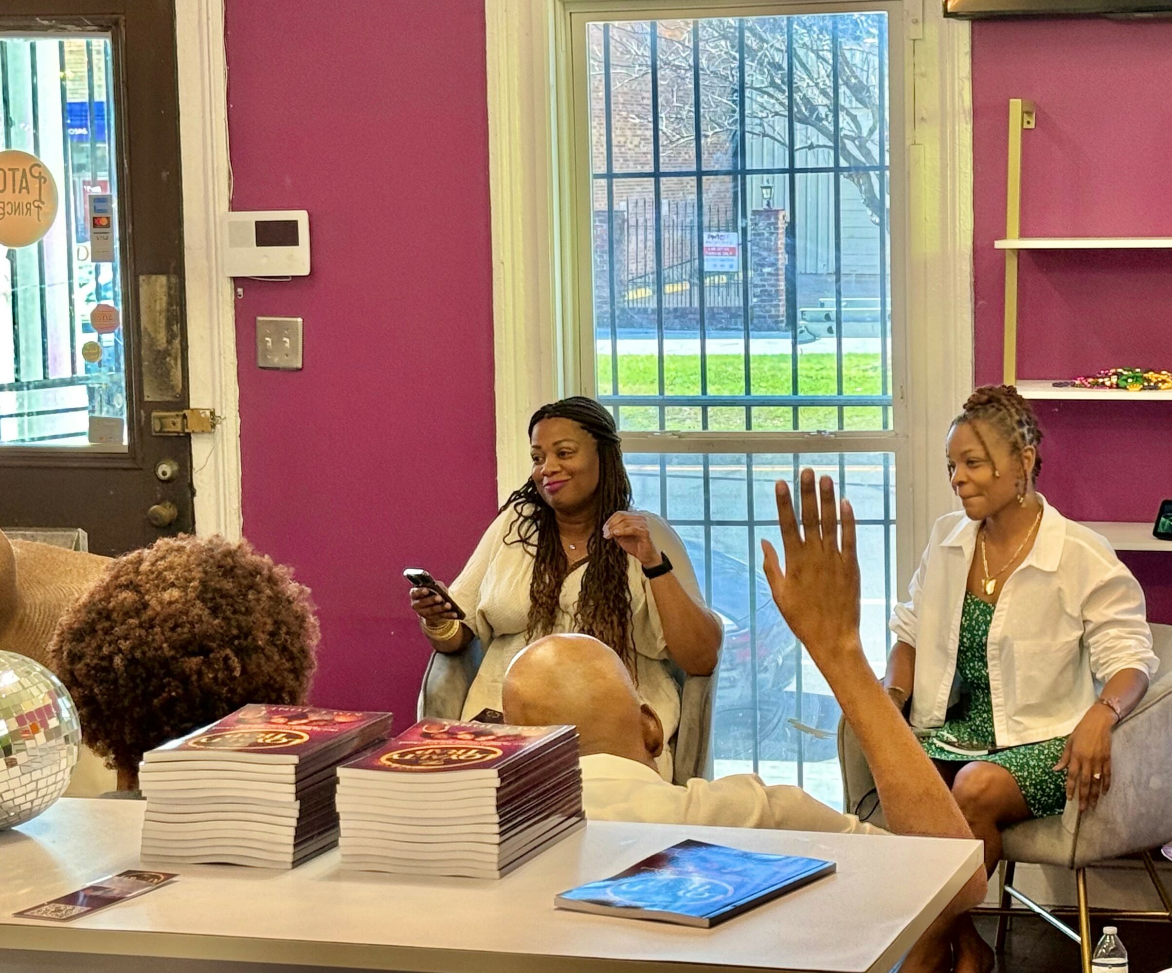 Two women seated in a colorful room with pink walls, large window, and shelves, engaging with an audience. One woman holds a phone.