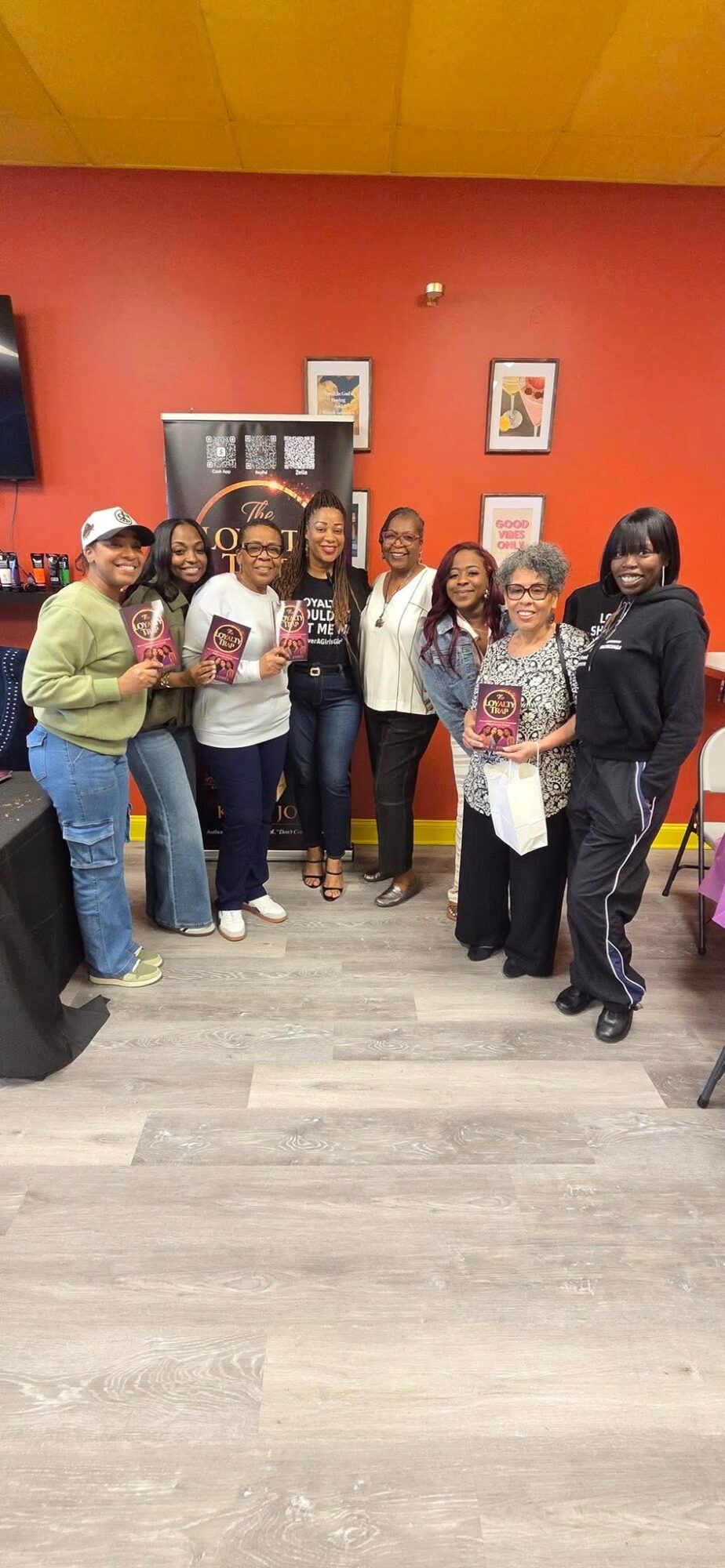 Group of women standing together indoors, some holding gift bags, in front of a red wall with framed pictures.