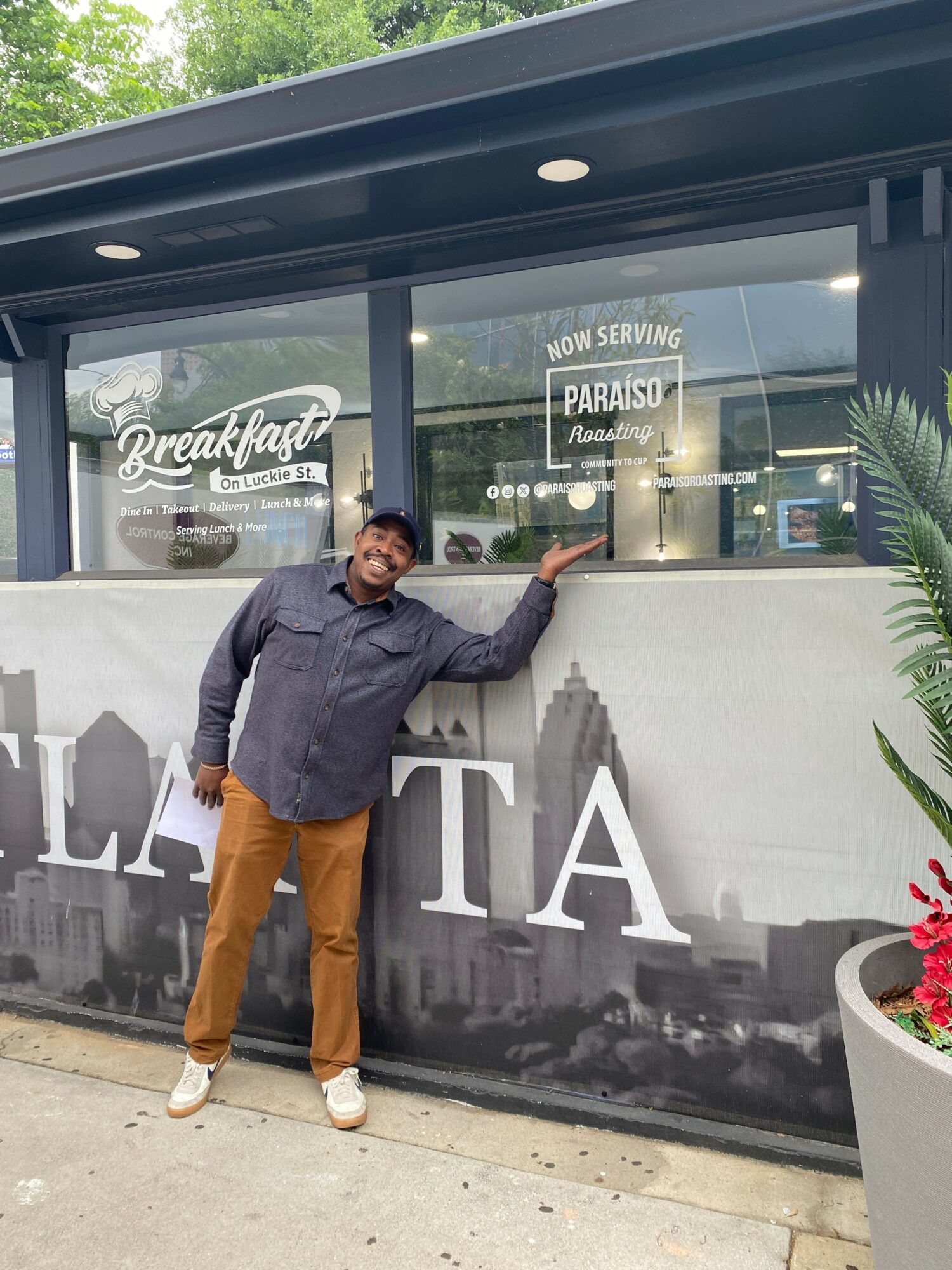 Person standing outside a restaurant, smiling, pointing at a sign in the window, with a cityscape mural on the wall behind.