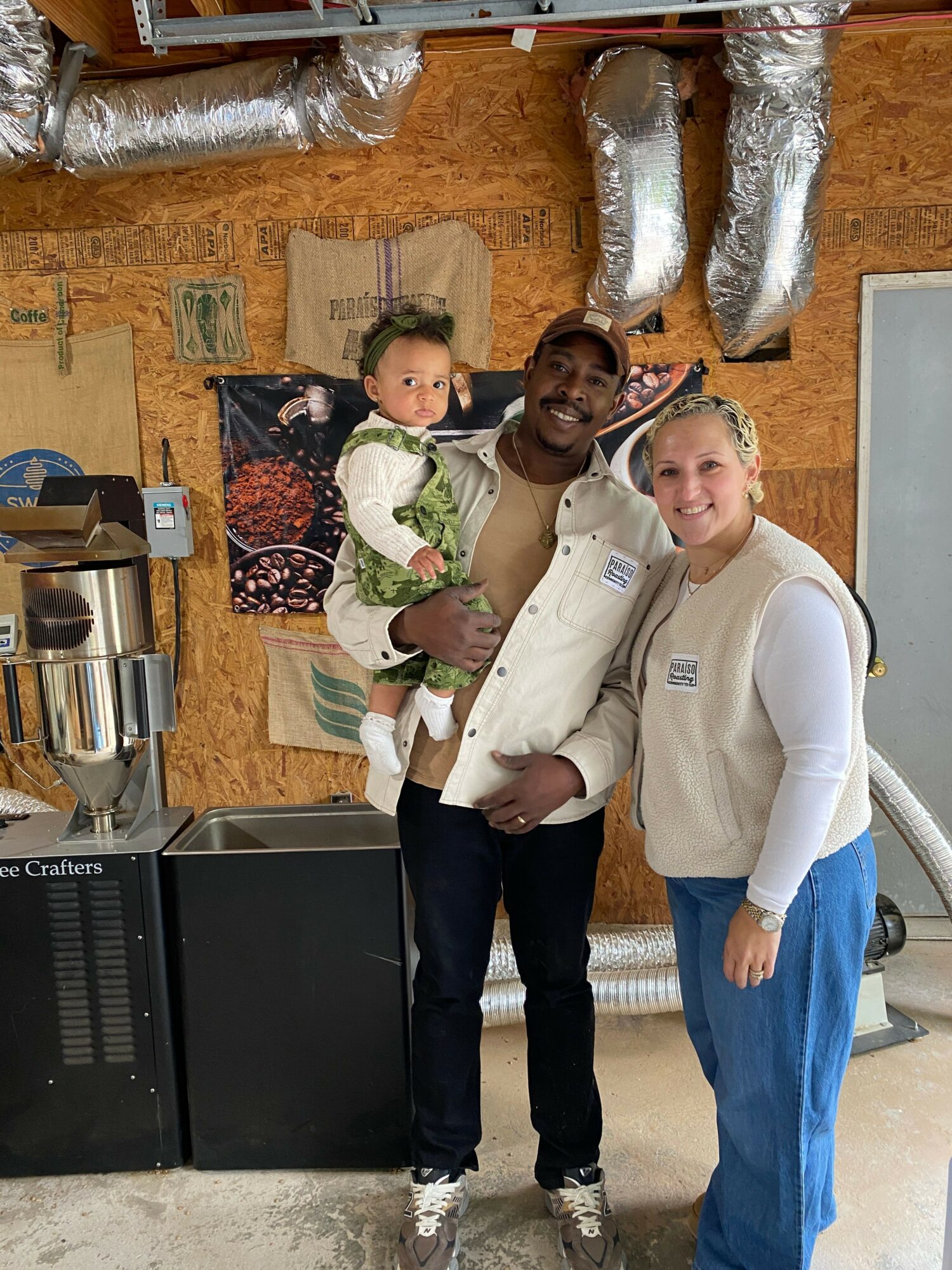 Three people standing indoors, one holding a child, with a wooden wall, metallic pipes, and equipment in the background.
