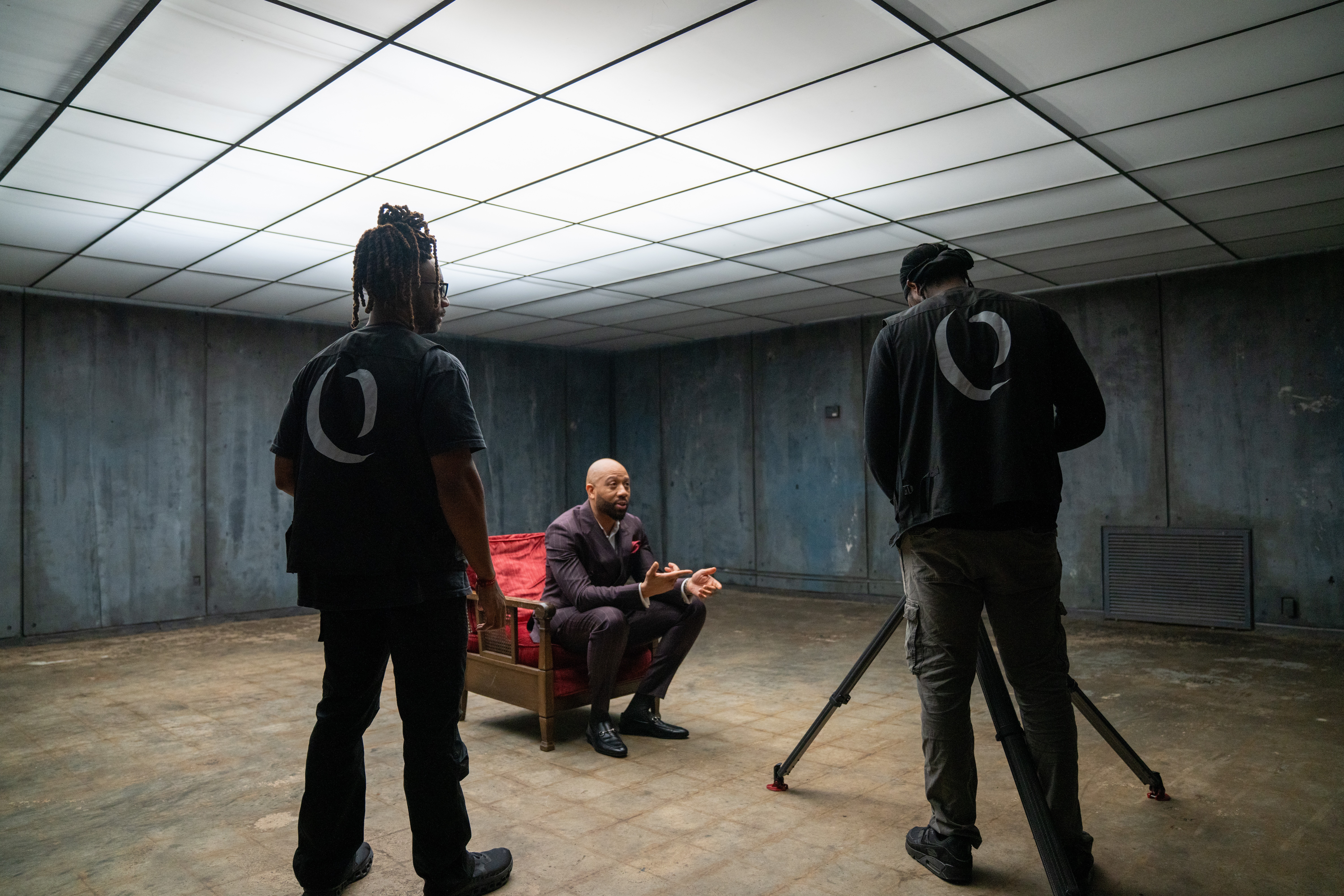 Two people stand in a room with a seated person using a phone, concrete walls, and a ceiling with illuminated panels.