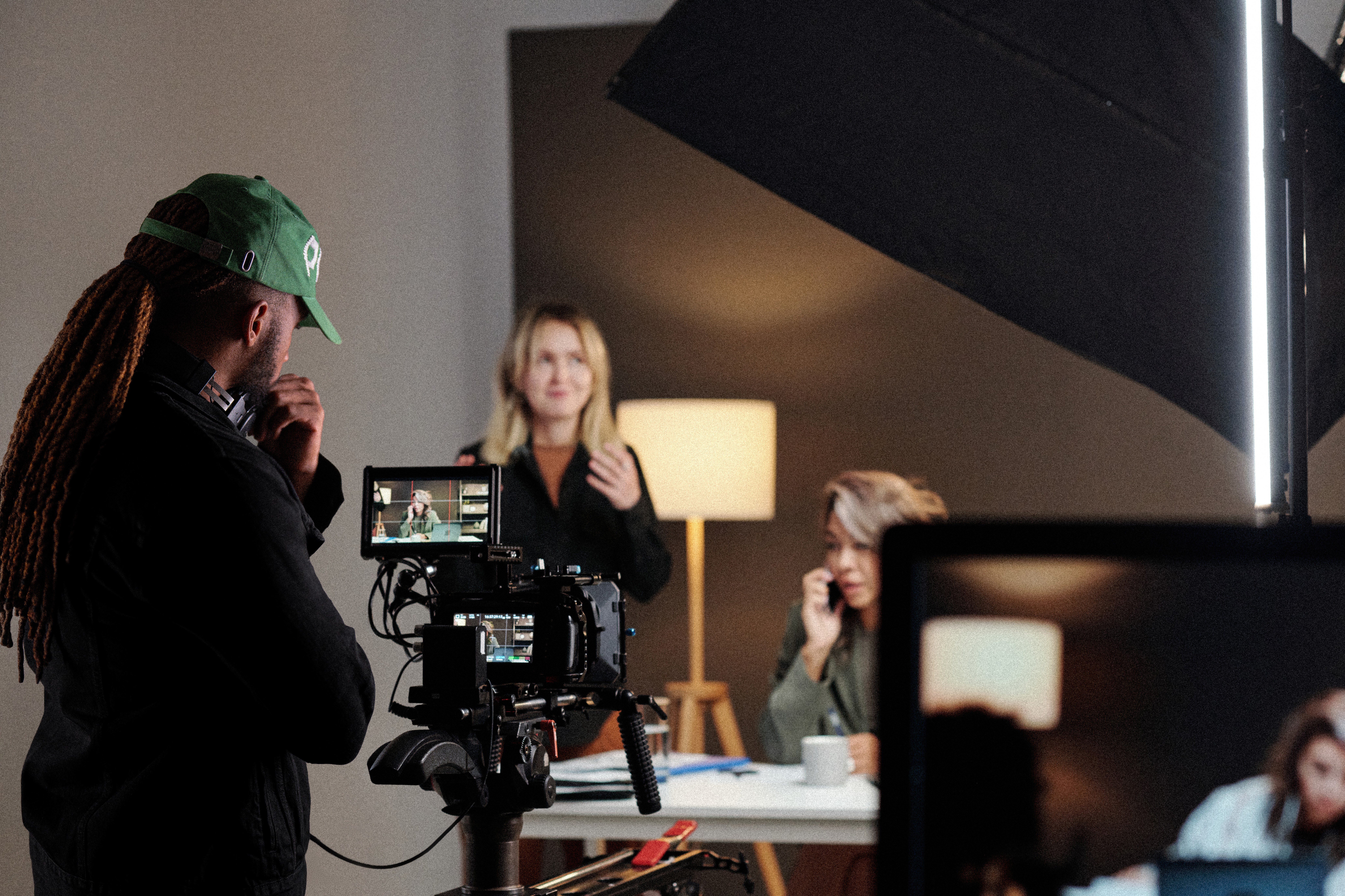 Person with long hair and green cap filming a woman speaking in a room with a lamp and a child, indoors.