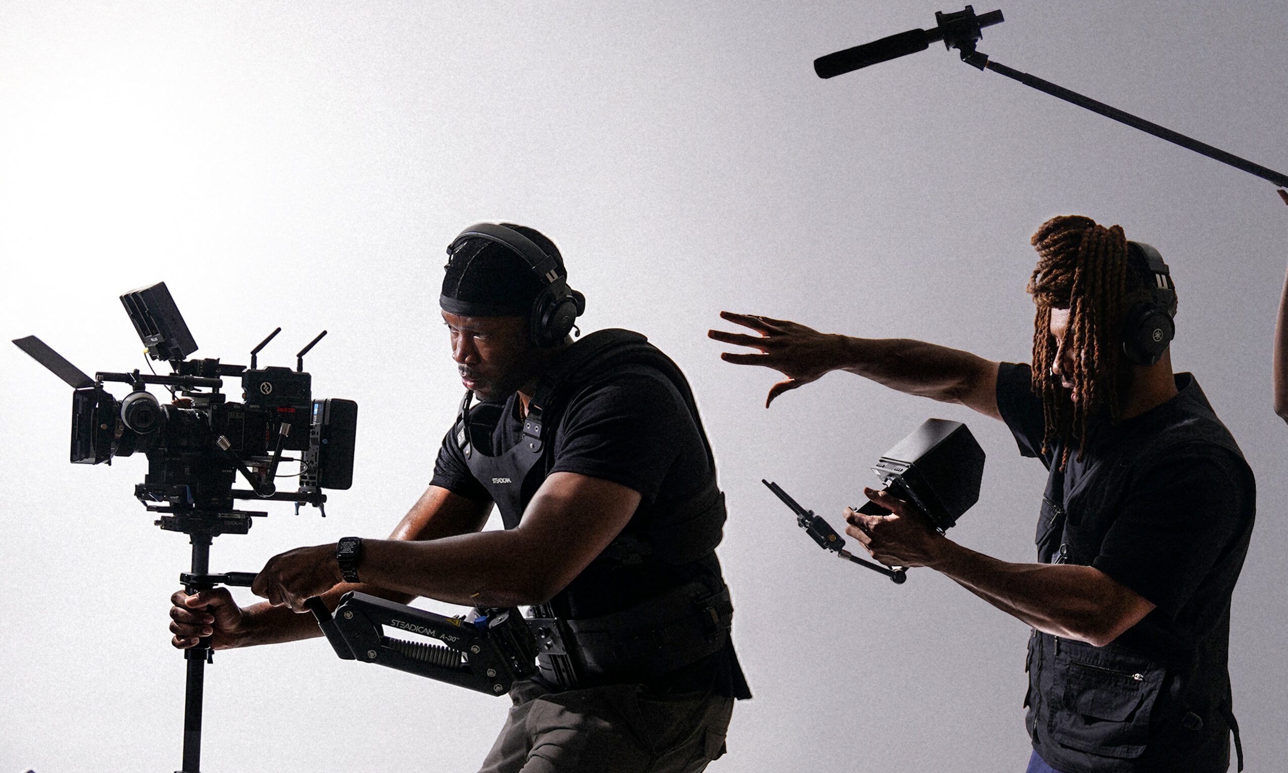 Two people working with filming equipment, one holding a camera and the other holding a phone, with a microphone overhead.