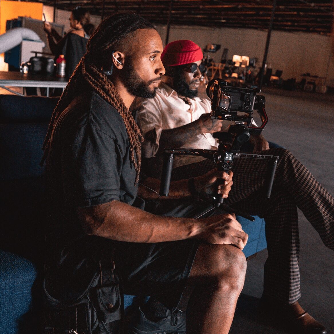 Two men sitting indoors, one with long dreadlocks and the other wearing a red hat, both focused on filming equipment.