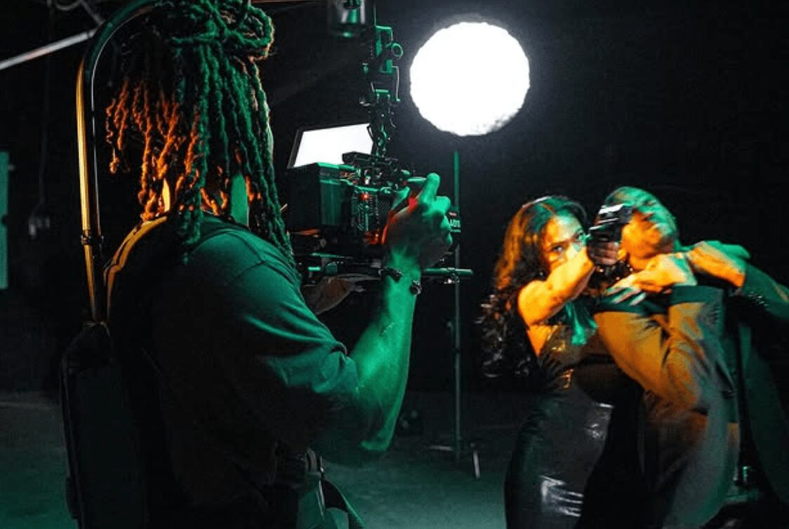 Two people in a dark studio, one with glasses and the other with long hair, appear to be in a conversation or performance.