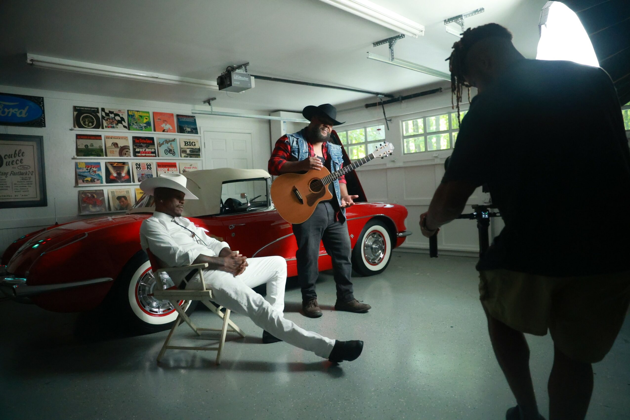 Person playing guitar and singing, another sitting on a chair, in a garage with a red car and posters on the wall.