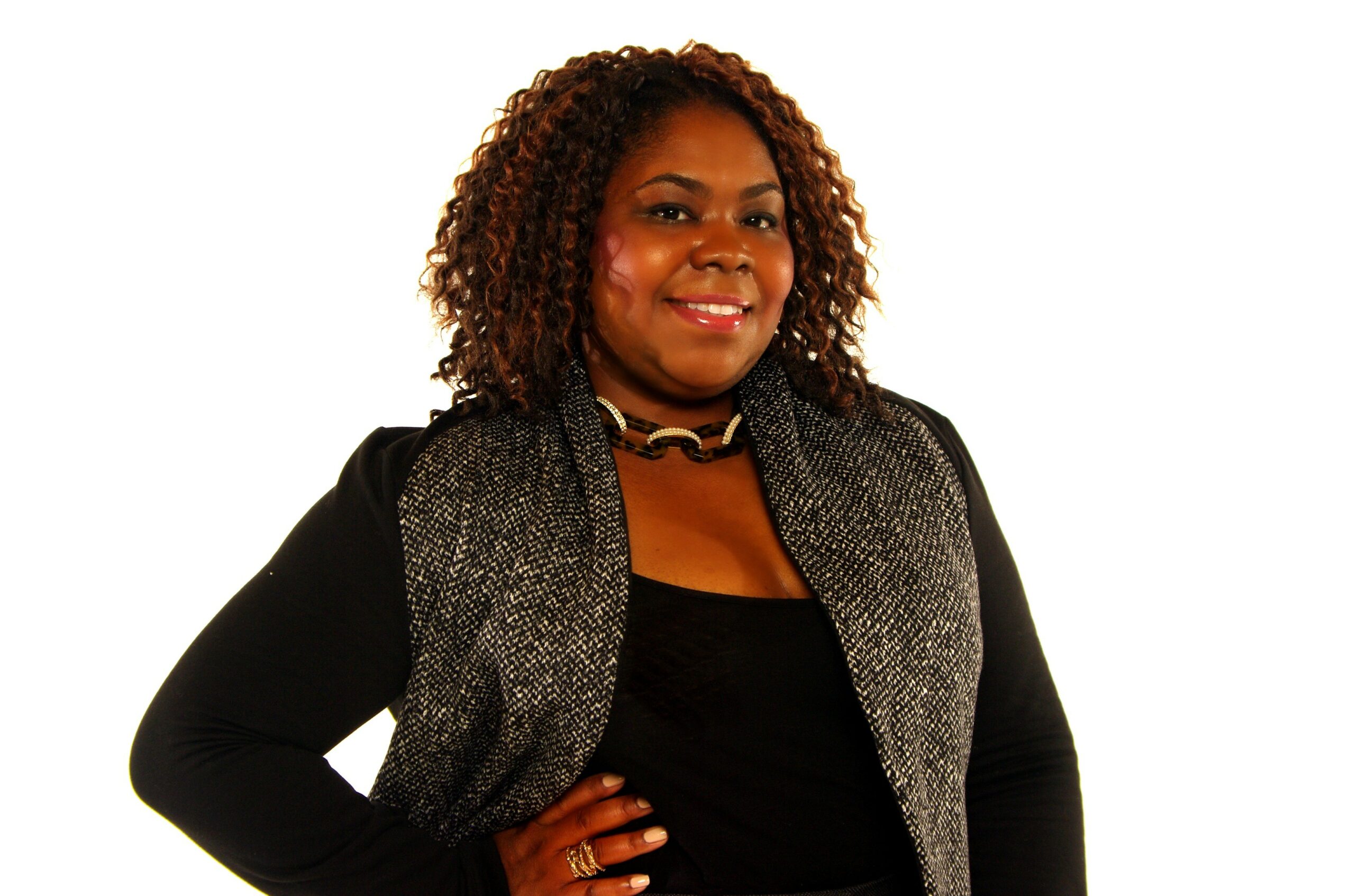 Woman with curly hair smiling, wearing a black top and gray vest, standing with hand on hip against white background.
