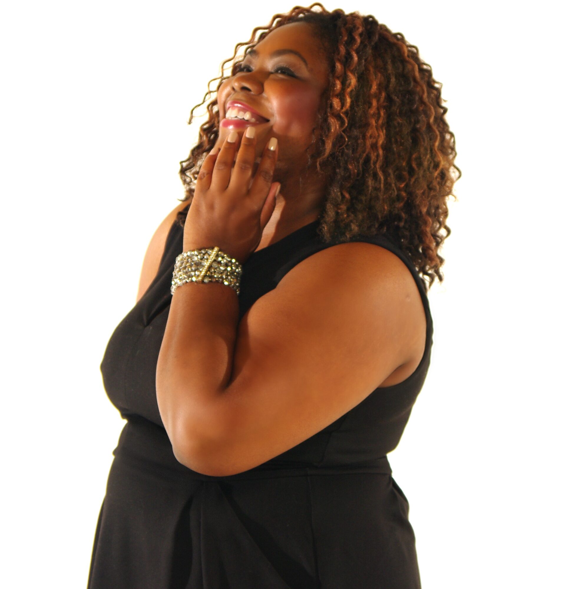 Smiling woman with curly hair wearing a black sleeveless top, touching her face, against a white background.