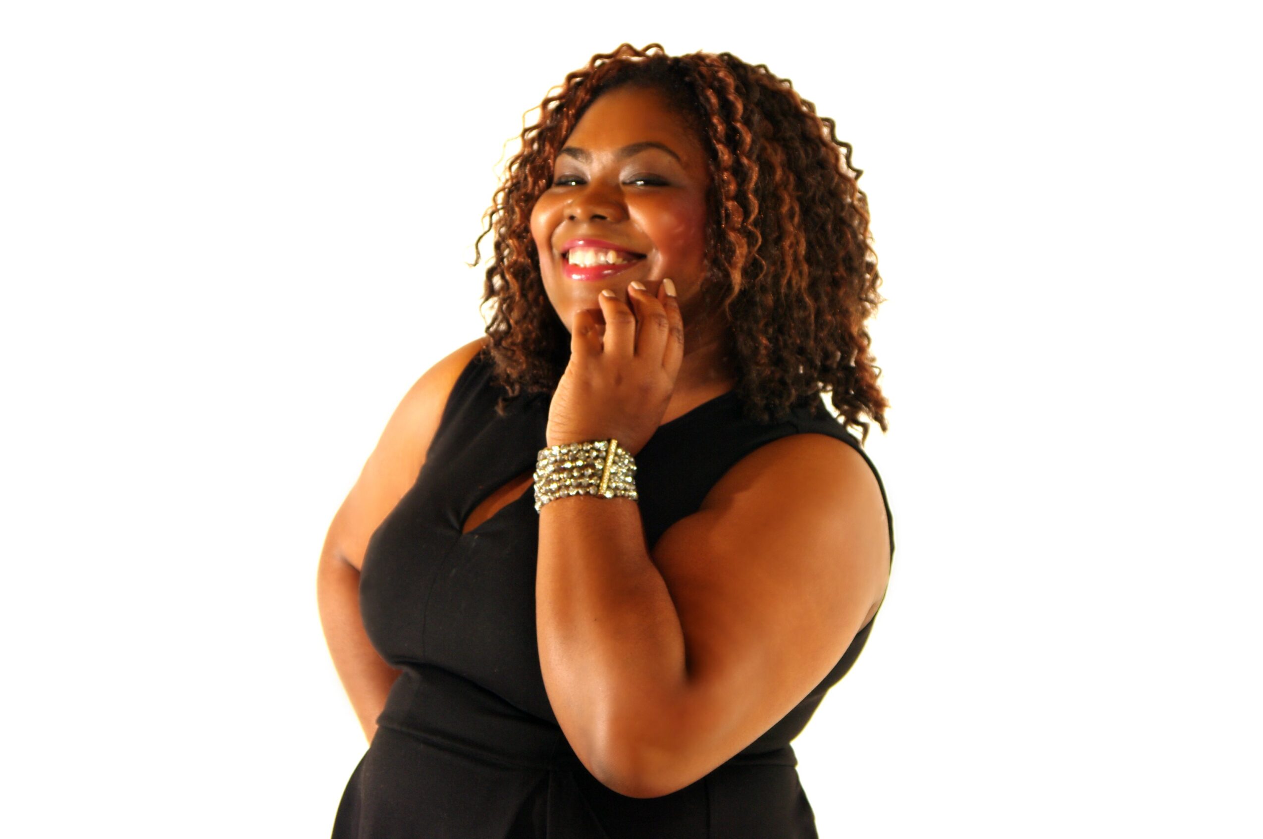 Smiling woman with curly hair wearing a black sleeveless dress and bracelet, touching her face with her hand, against a white background.