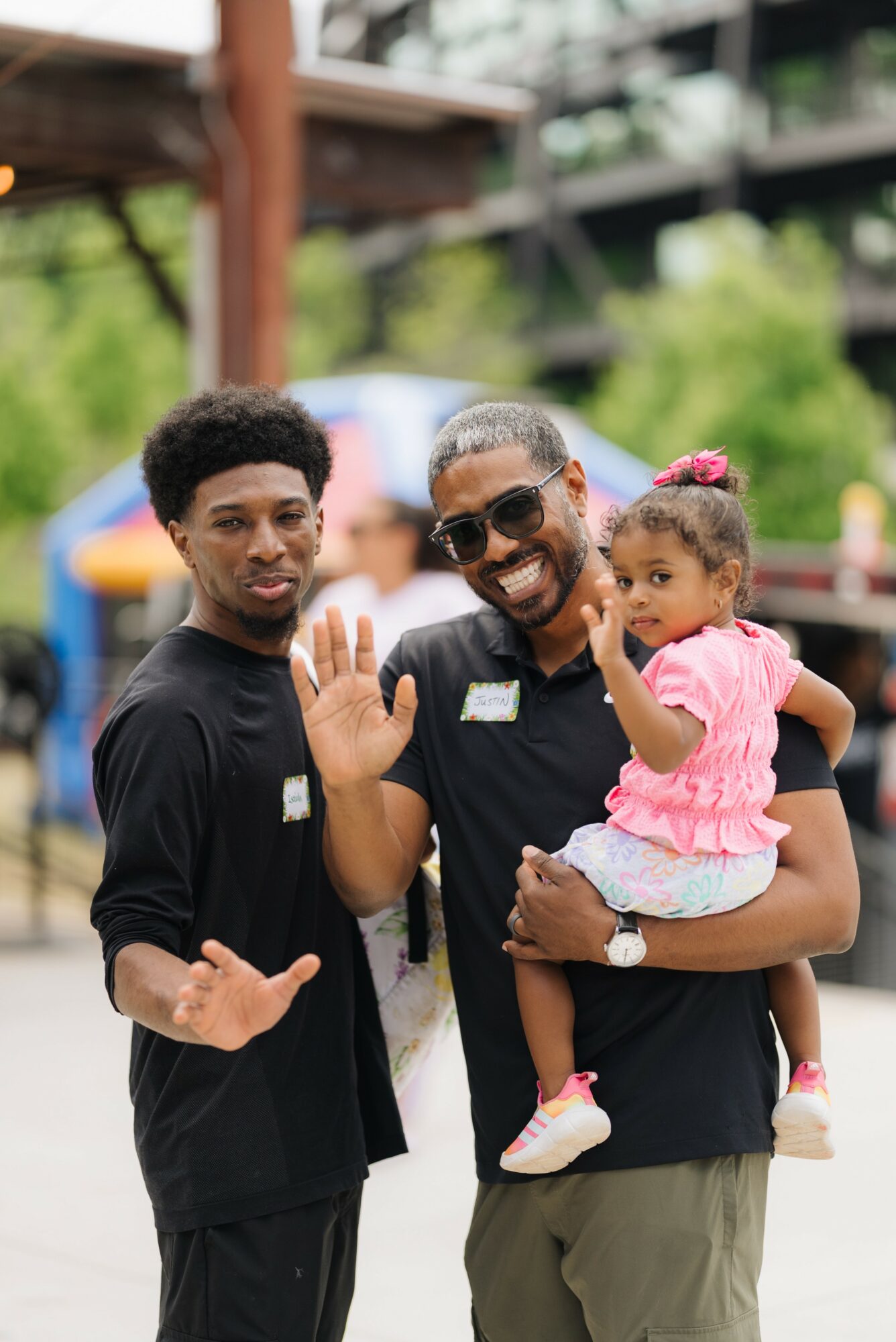 Two men and a young girl outdoors, one man holding the girl, all smiling and waving, background with trees and structures.