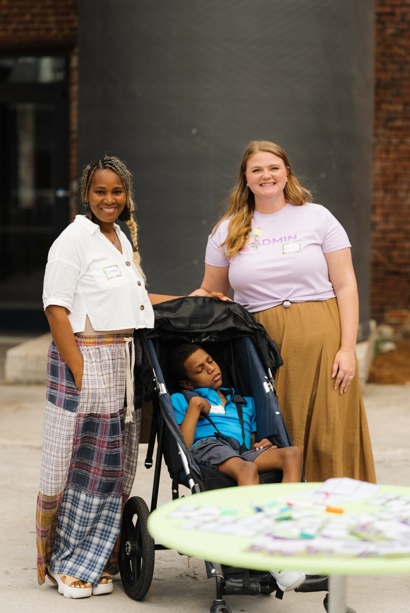 Two women standing outdoors with a child in a stroller, smiling, with a table in the foreground.