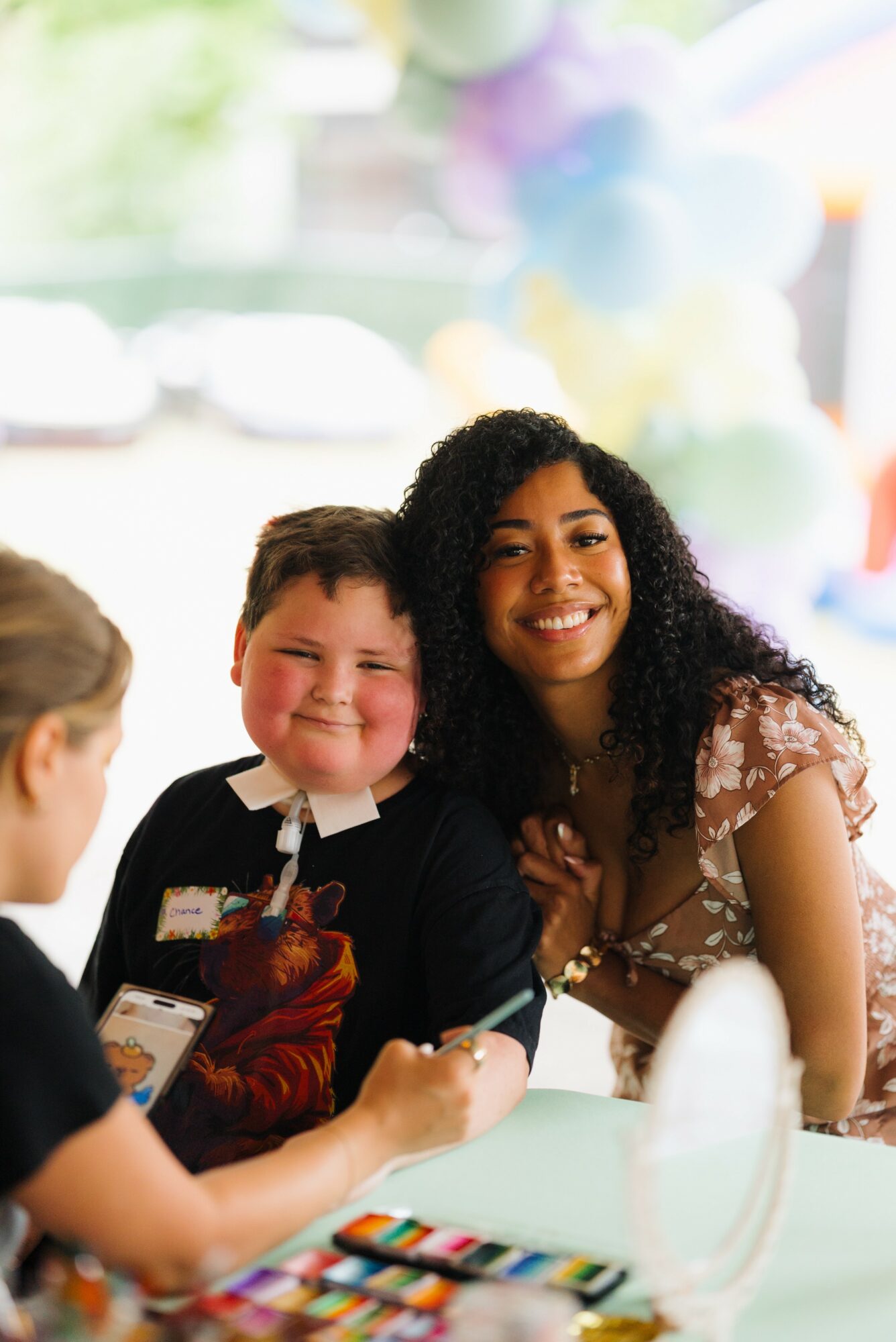 Smiling young boy and woman with curly hair sitting at a table with colorful markers, in a bright room with balloons.