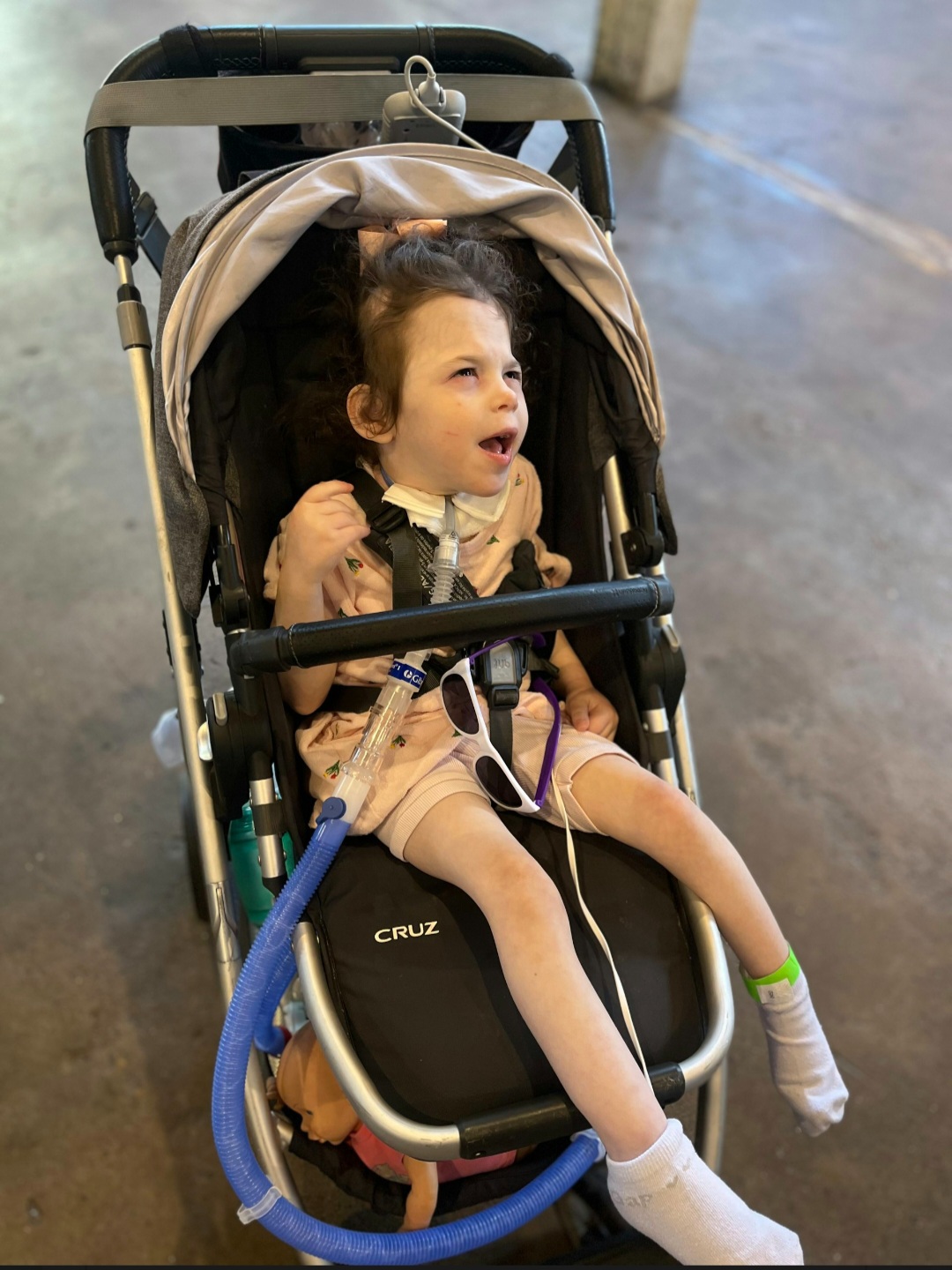 Young child sitting in a stroller, smiling and looking to the side, with a blue tube connected to the stroller.
