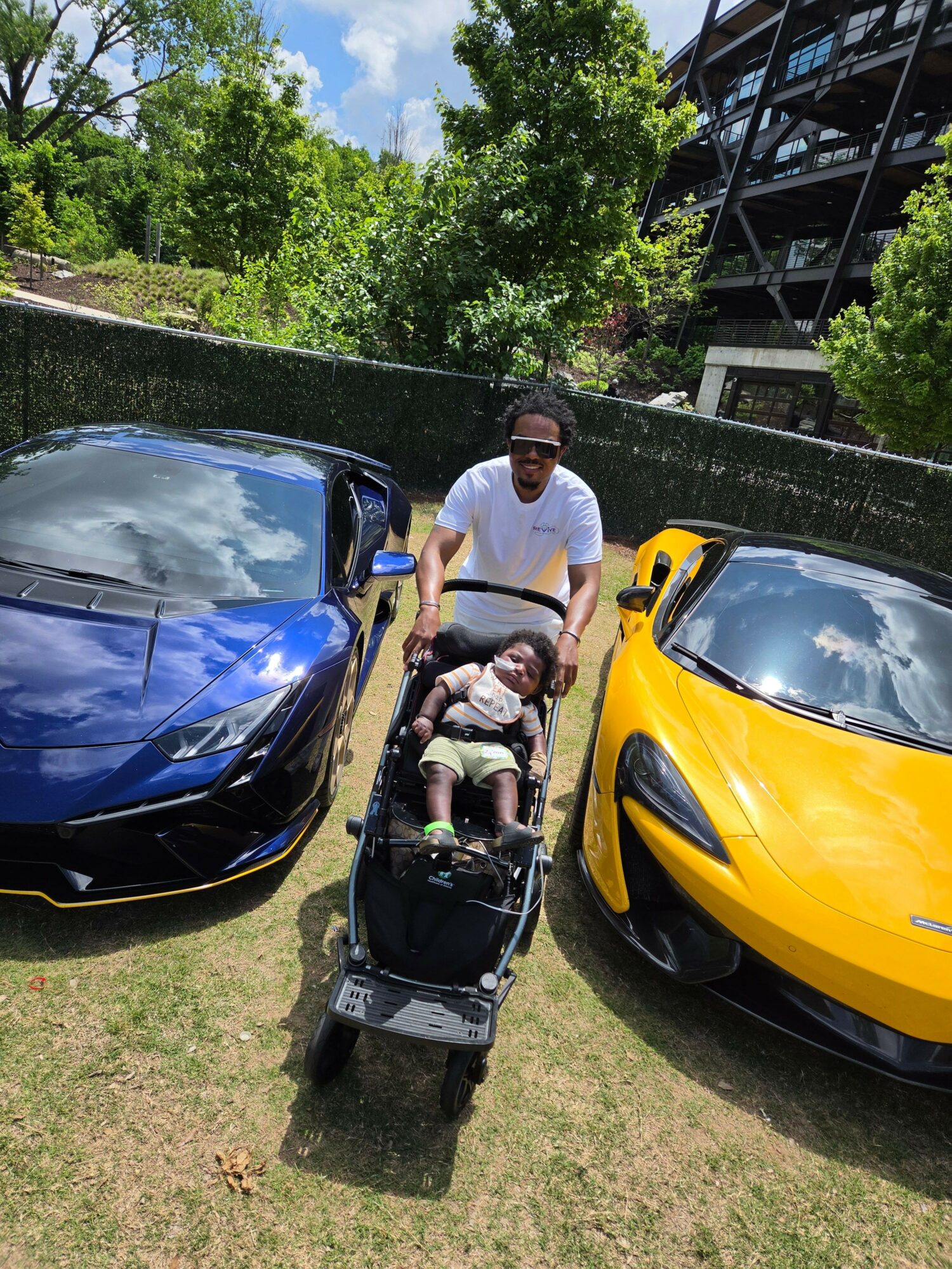 Woman with a stroller and a child walking beside two parked sports cars, one yellow and one blue, outdoors on grass.