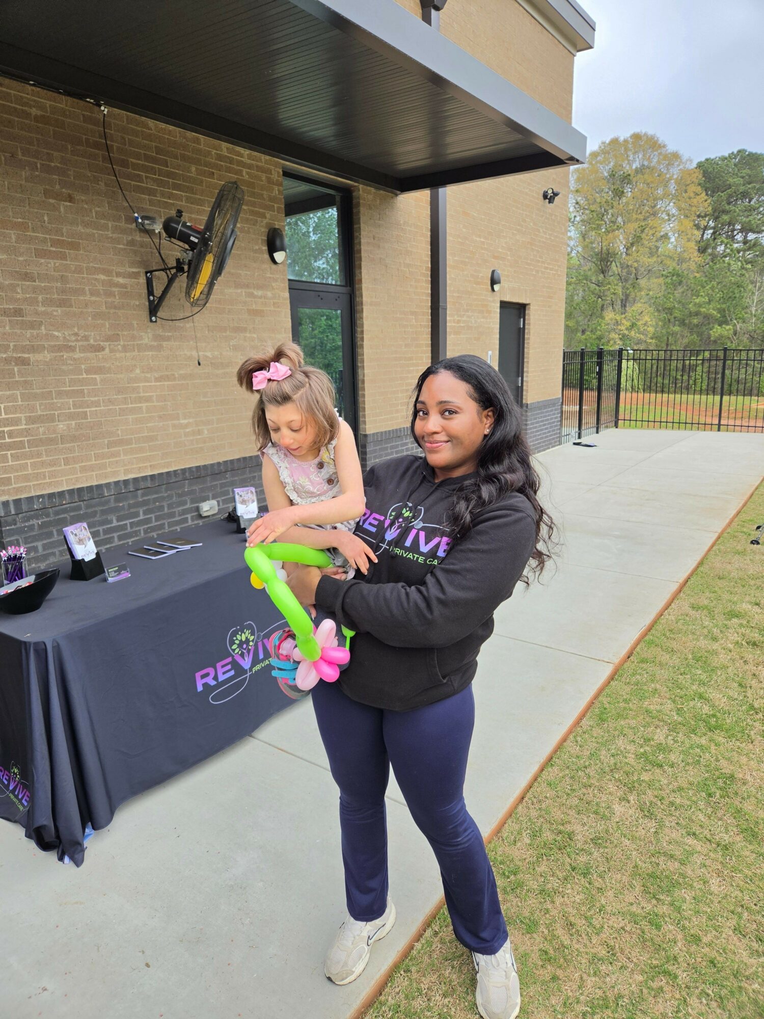 Woman and child with balloon animals outside near a table, with a bicycle and building in background.
