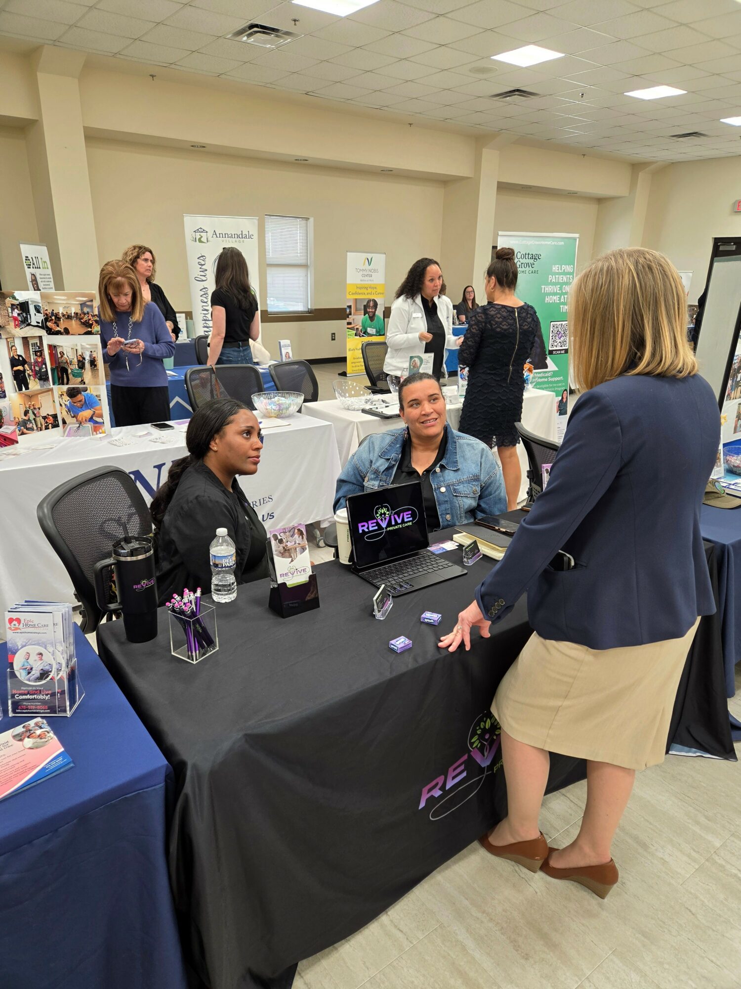 Three women engaged in conversation at a table in a conference room, with other people and booths in the background.