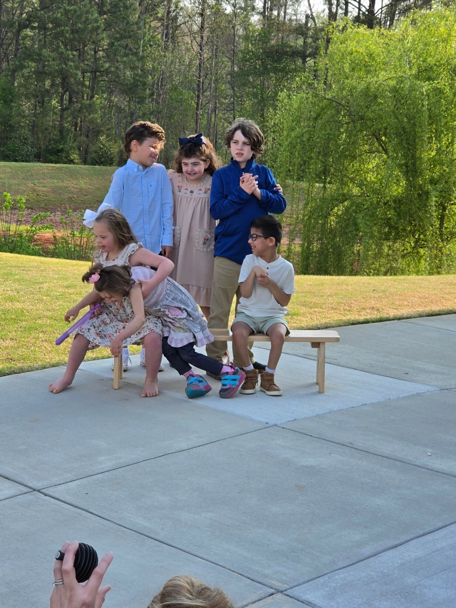 Children and an adult walking outdoors on a paved path, surrounded by trees and grass, with some children holding hands.