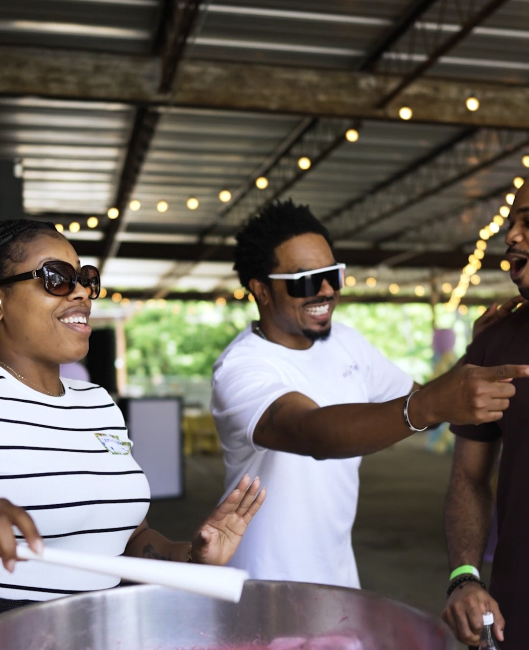 Three people smiling and talking under a metal roof with string lights, one person pointing, outdoors setting.