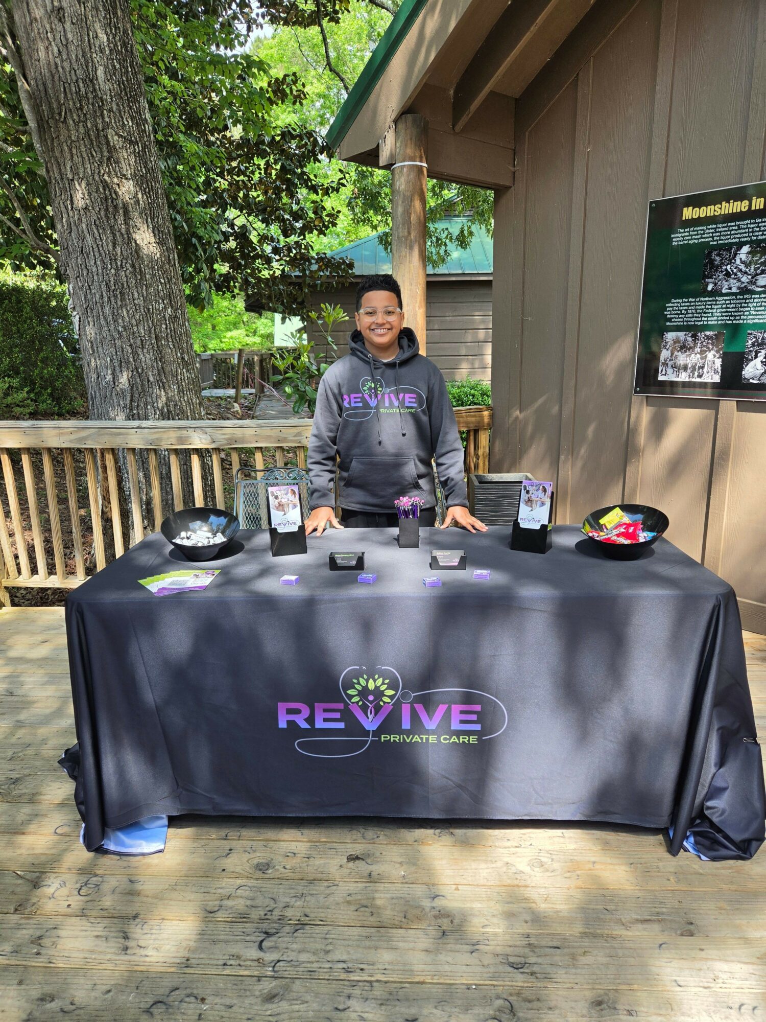Person standing behind a table with promotional materials outdoors, trees and a wooden fence in background.