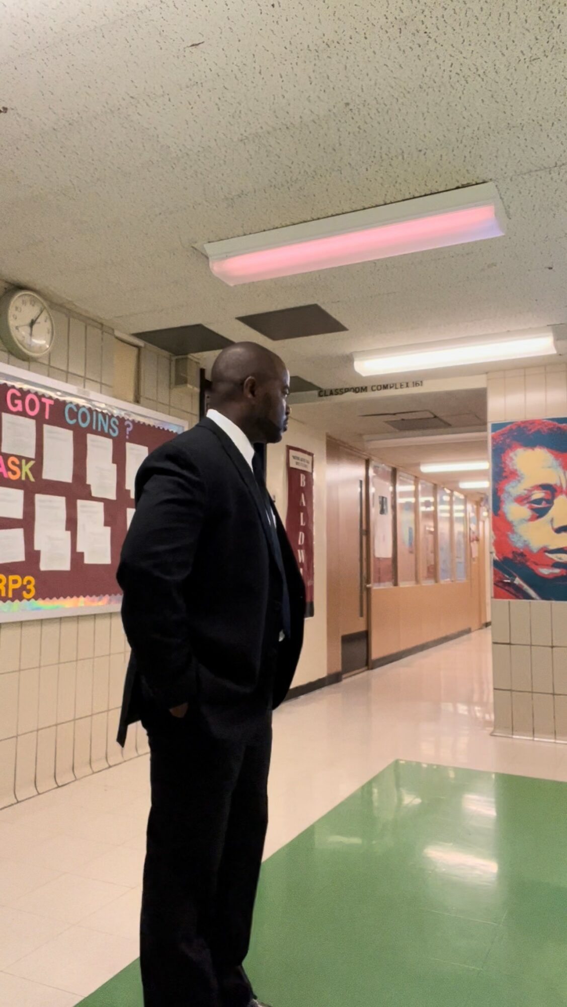 Man in suit standing in school hallway with bulletin boards and a portrait of a woman on the wall.