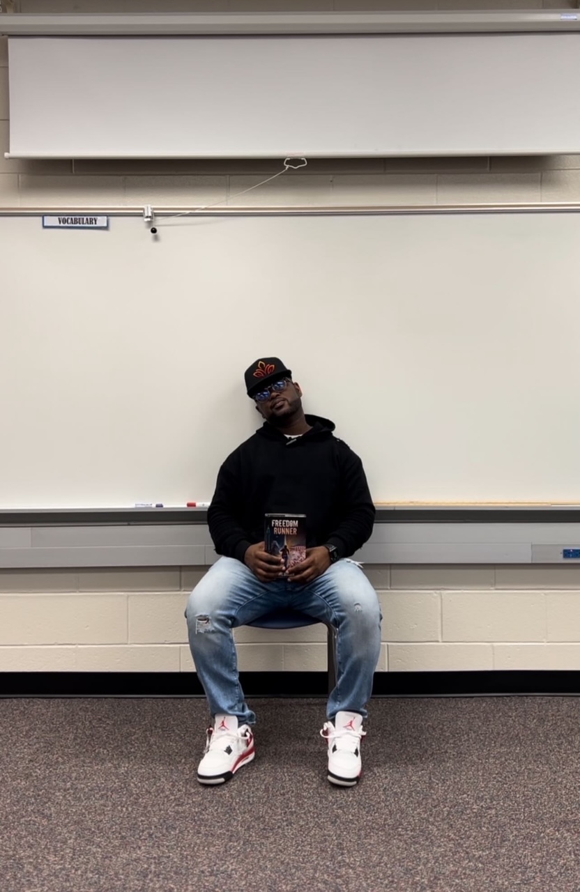 Person sitting on a chair in front of a whiteboard, holding a phone, wearing a black hoodie, jeans, and white sneakers.