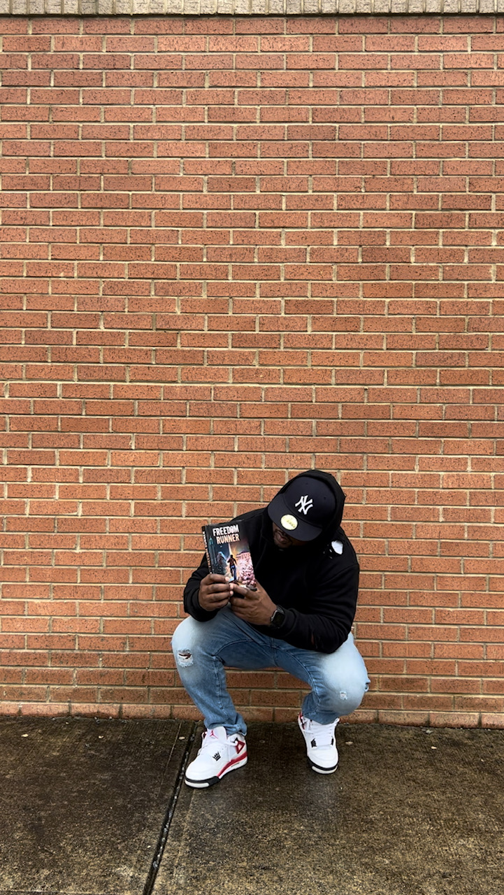 Person crouching in front of a brick wall, wearing a black hoodie, white sneakers, and a black cap, holding a phone.