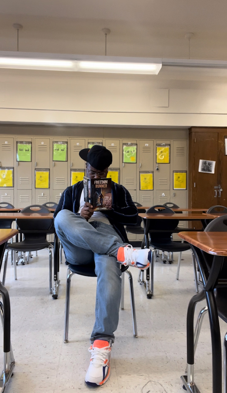 Person sitting on a chair in a classroom, taking a selfie with a phone, with lockers and tables in the background.