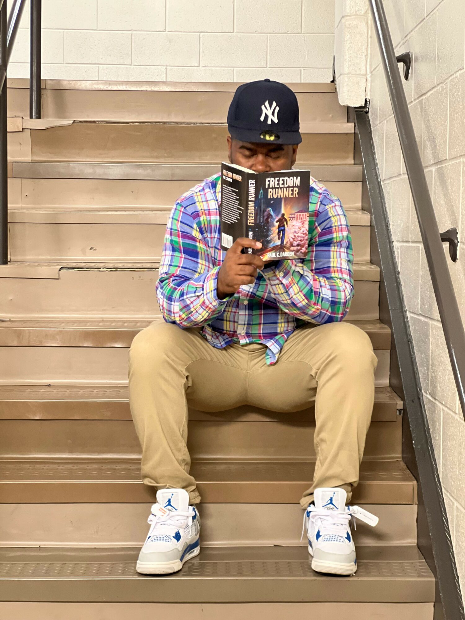 Person sitting on stairs, wearing a cap, plaid shirt, beige pants, and white sneakers, holding a magazine and taking a selfie.