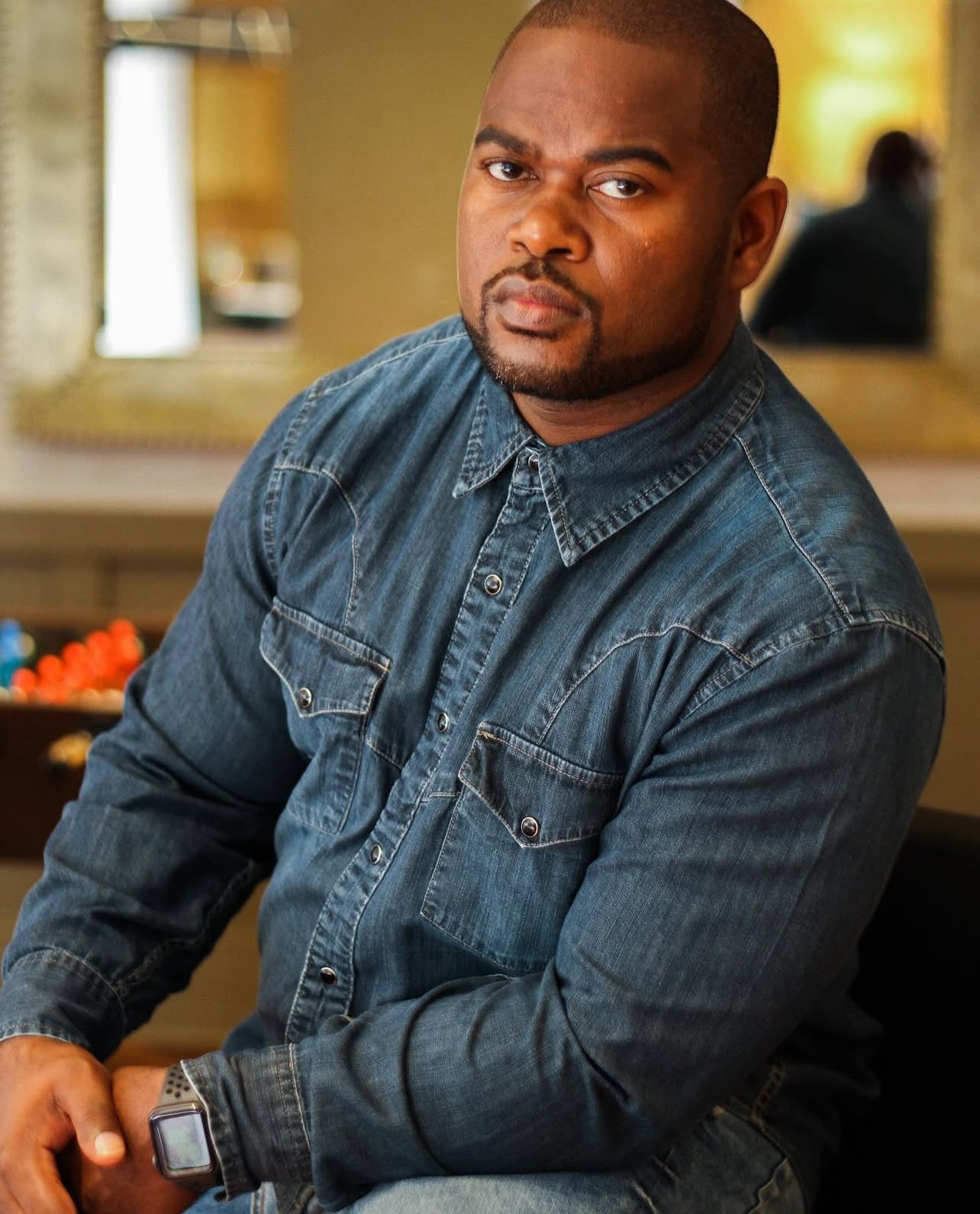 Man with short hair and a beard wearing a denim shirt, sitting indoors with a neutral expression.